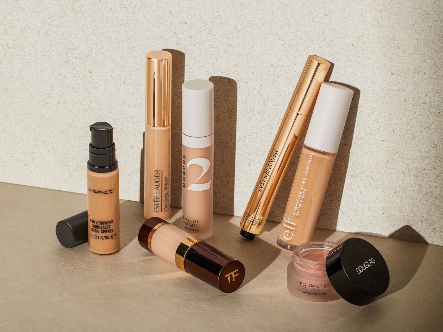 Eine Auswahl an Concealern von Top-Marken wie MAC, Estée Lauder, Morphe, Yves Saint Laurent, elf und Douglas, präsentiert in verschiedenen Flaschen und Tuben, die auf einem hellen Hintergrund stehen.