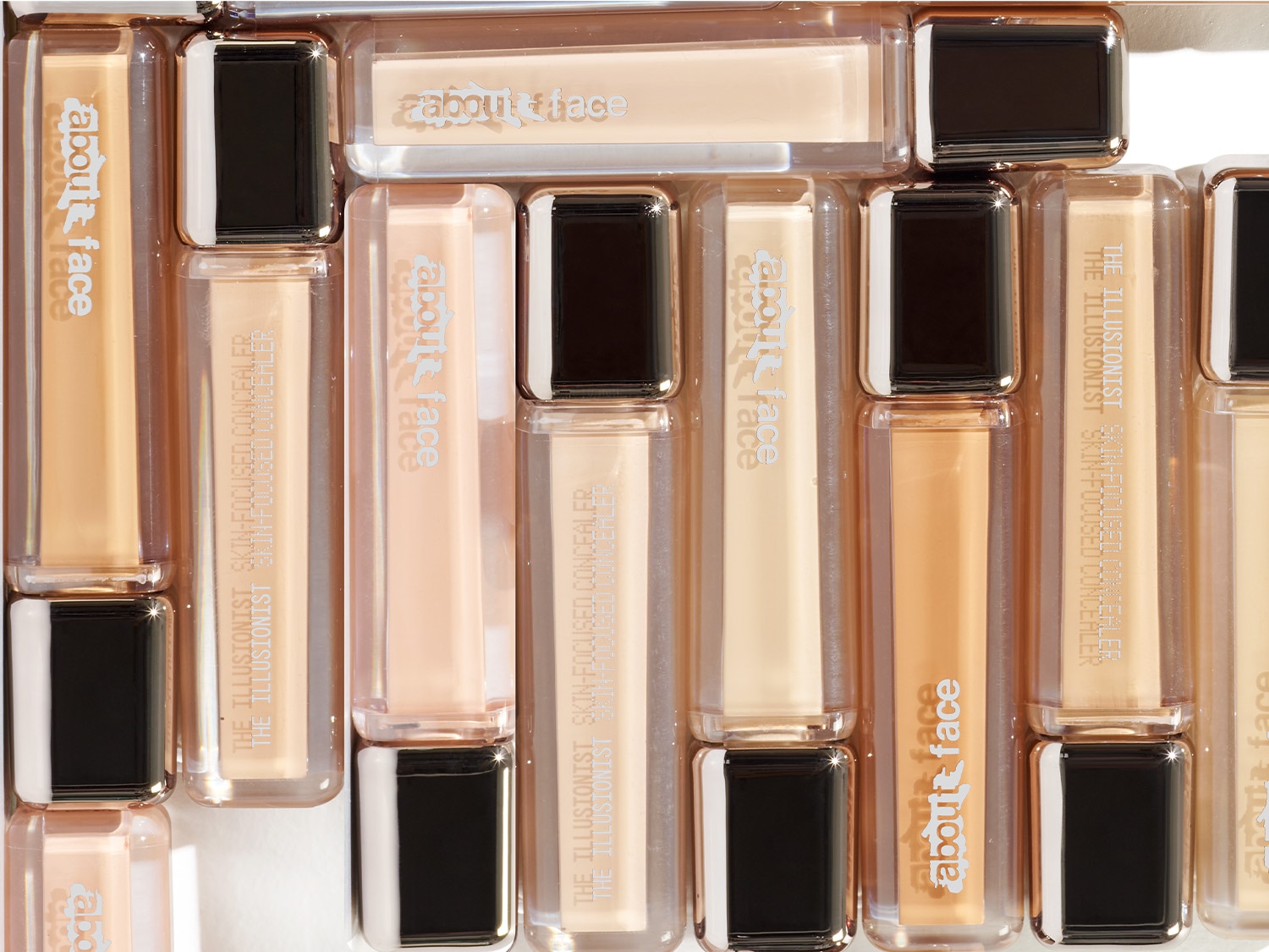 Šiame vaizde matyti „About Face The Illusionist Skin-Focused Concealer“ maskuokliai, išdėstyti eilėmis, su juodais dangteliais ir permatomais buteliukais, atskleidžiančiais įvairius odos atspalvius.
