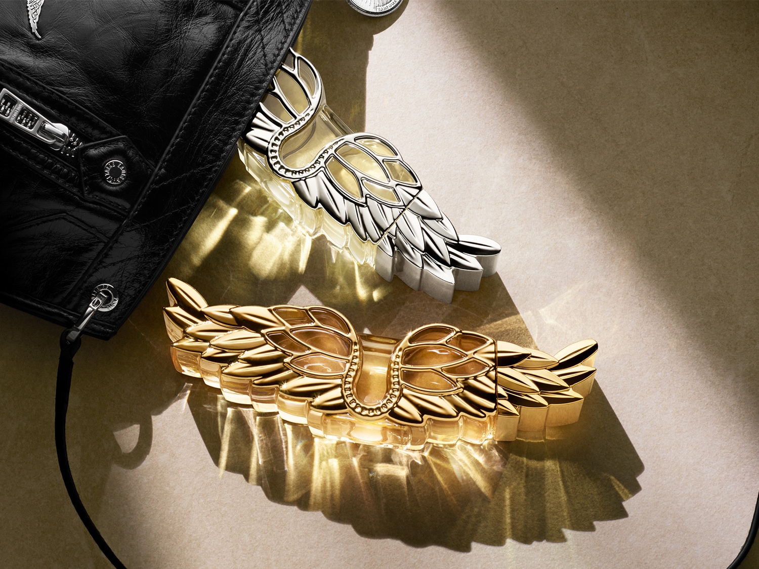 Nahaufnahme von zwei Parfümflakons in Flügelform, eine goldene und eine silberne, neben einer schwarzen Lederjacke.