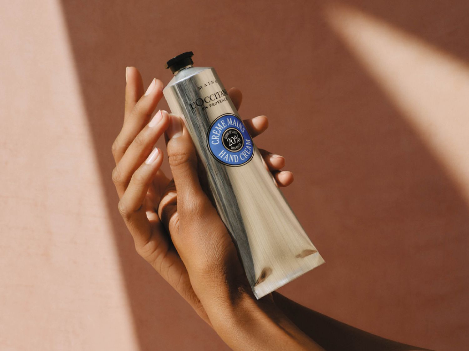 Eine Hand hält eine silberne Tube Handcreme von L'OCCITANE mit blauem Etikett, das 20 % Sheabutter hervorhebt, vor einem sanften, pfirsichfarbenen Hintergrund.