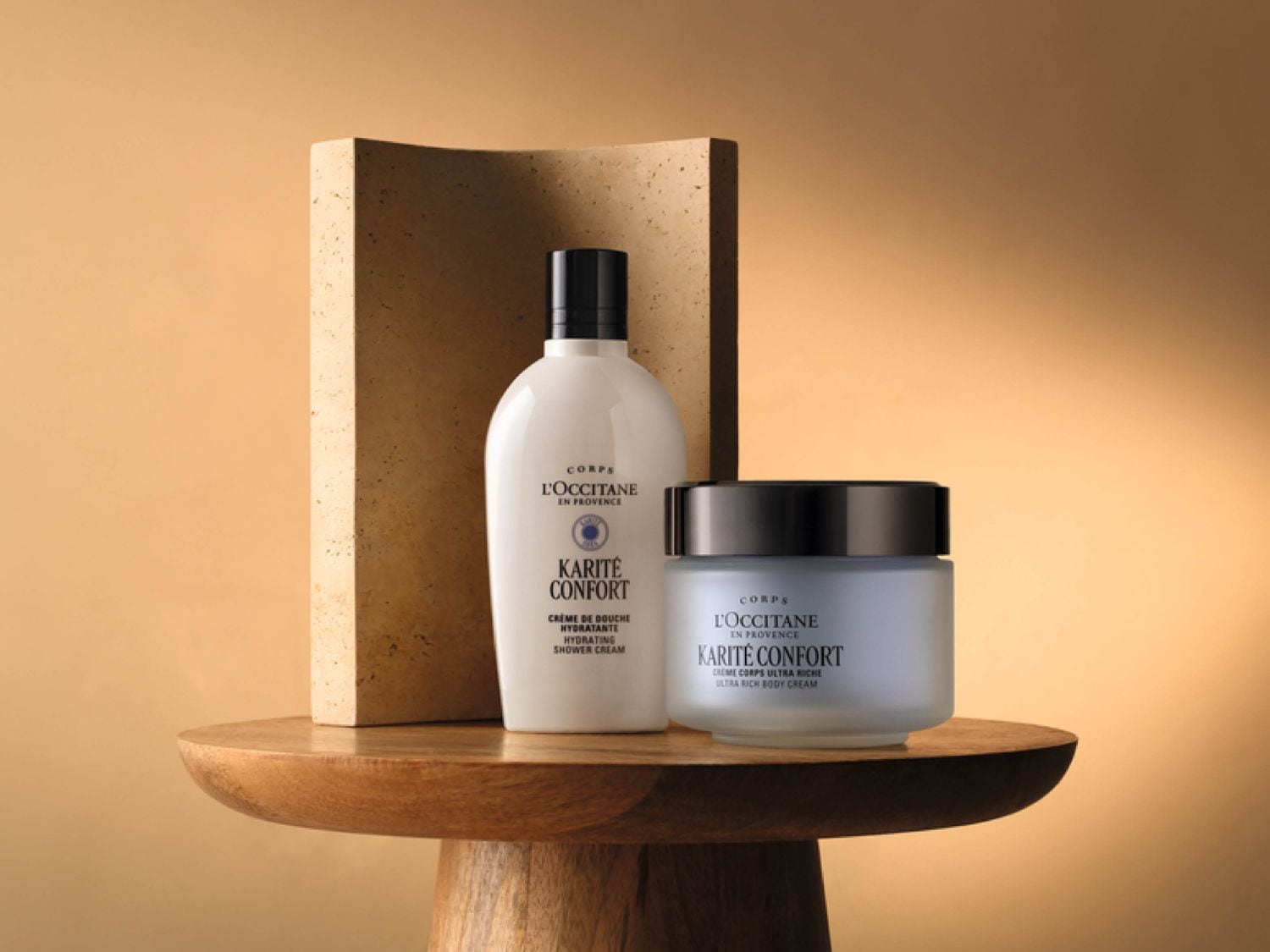 Auf einem Holztisch stehen eine weiße Flasche Karité Duschcreme und ein blassblauer Tiegel Karité Körpercreme von L'OCCITANE vor einem beigen Hintergrund.