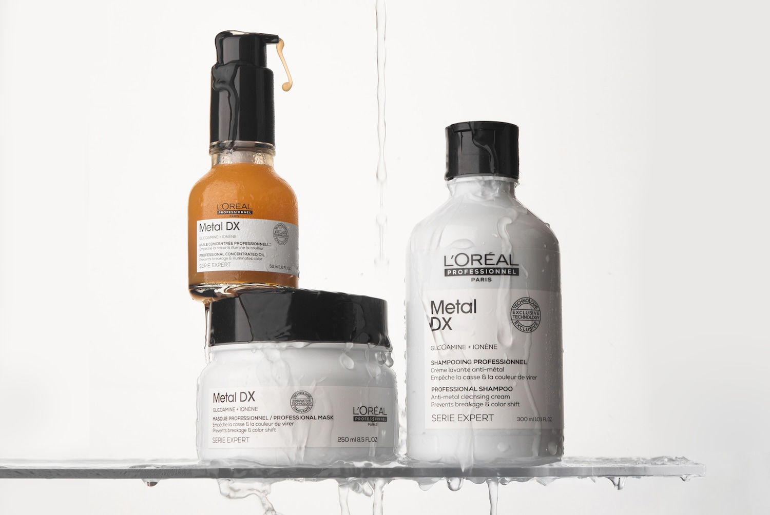 Drei nasse, mit Wassertropfen bedeckte L'Oréal Professionnel Metal DX Haarpflegeprodukte stehen auf einer nassen Glasplatte vor hellem Hintergrund: Links oben das goldene Haaröl, darunter die weiße Maske und rechts das weiße Shampoo, während im Hintergrund ein dünner Wasserstrahl herabfließt.