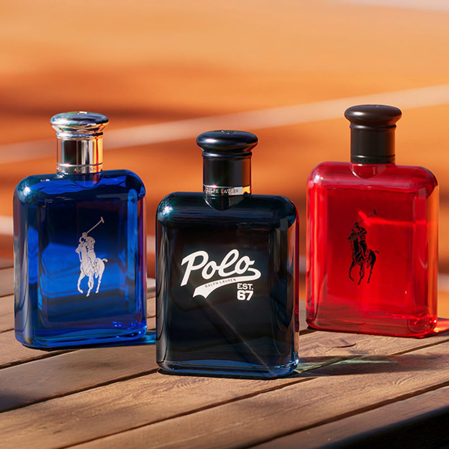 Drei Ralph Lauren Polo Parfümflaschen in Blau, Schwarz und Rot stehen auf einem Holztisch vor einem orangefarbenen Hintergrund.