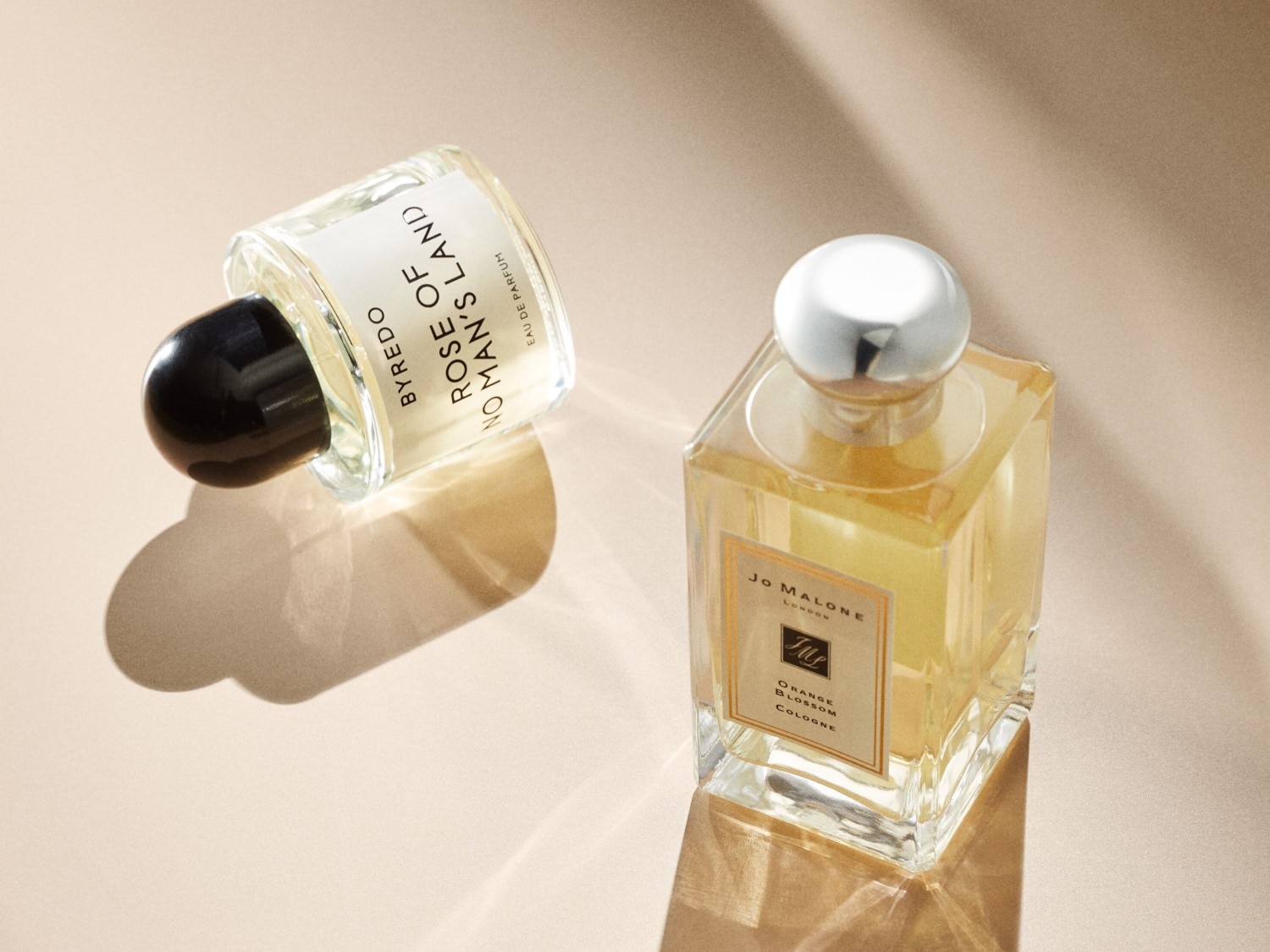 Twee parfumflessen, een van Byredo Rose of No Man's Land met een zwarte dop en een van Jo Malone Orange Blossom Cologne met een zilveren dop, liggen op een beige achtergrond.