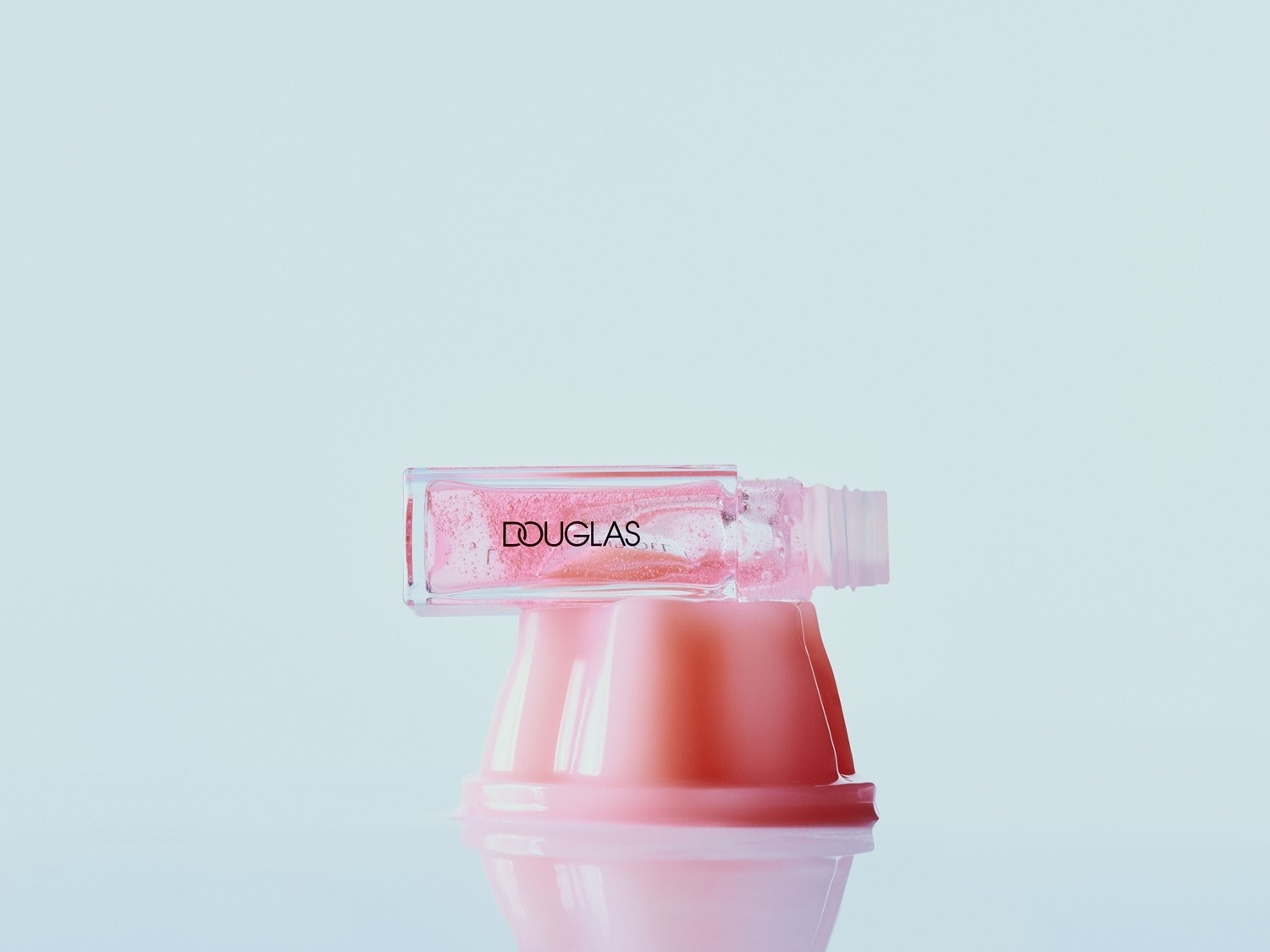 Ein Douglas Lipgloss in einem transparenten, rechteckigen Fläschchen mit schwarzem Logo, gefüllt mit schimmerndem, rosafarbenem Gloss, liegt auf einem rosafarbenen, pilzförmigen Ständer.