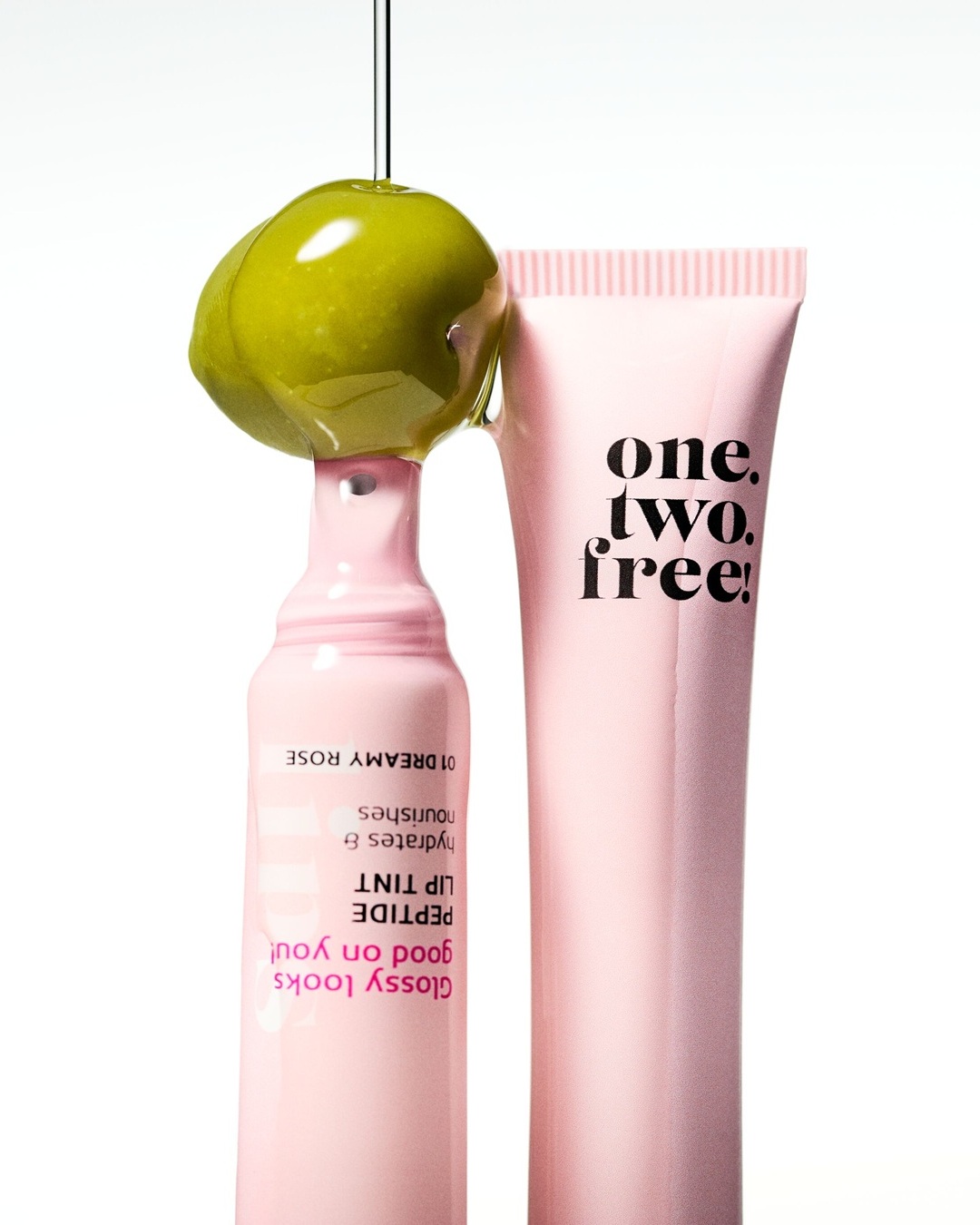 Das Bild zeigt zwei rosafarbene Tuben von "one.two.free!" mit der Aufschrift "PEPTIDE LIP TINT 01 DREAMY ROSE", die glänzende Lippen, Feuchtigkeit und Pflege versprechen, während eine grüne Olive mit Öl darüber schwebt.