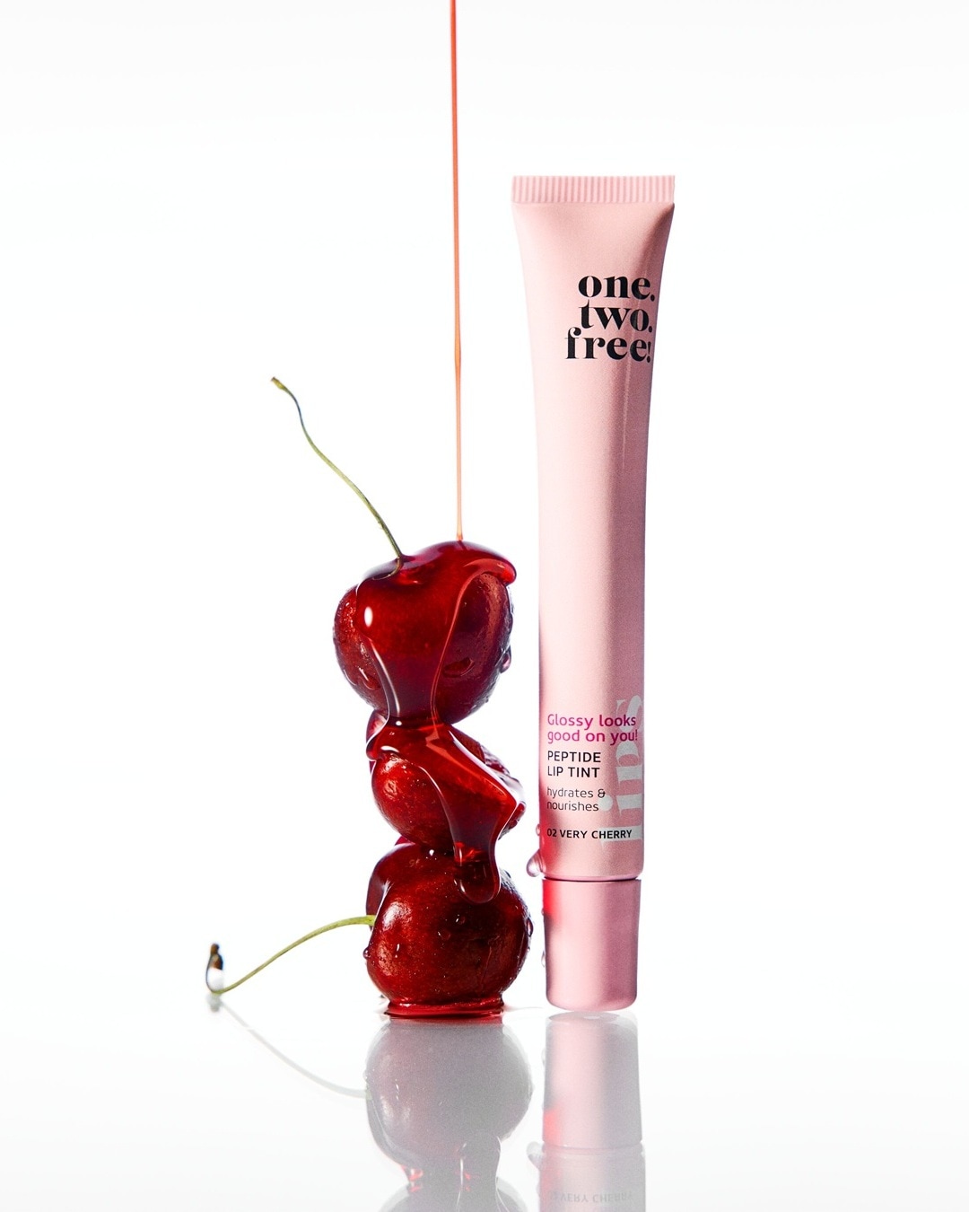 Das One.Two.Free Peptide Lip Tint in der Farbe "Very Cherry" ist ein feuchtigkeitsspendender und pflegender Lipgloss, der auf dem Bild neben drei glänzenden Kirschen steht, die mit einer roten Flüssigkeit überzogen sind.
