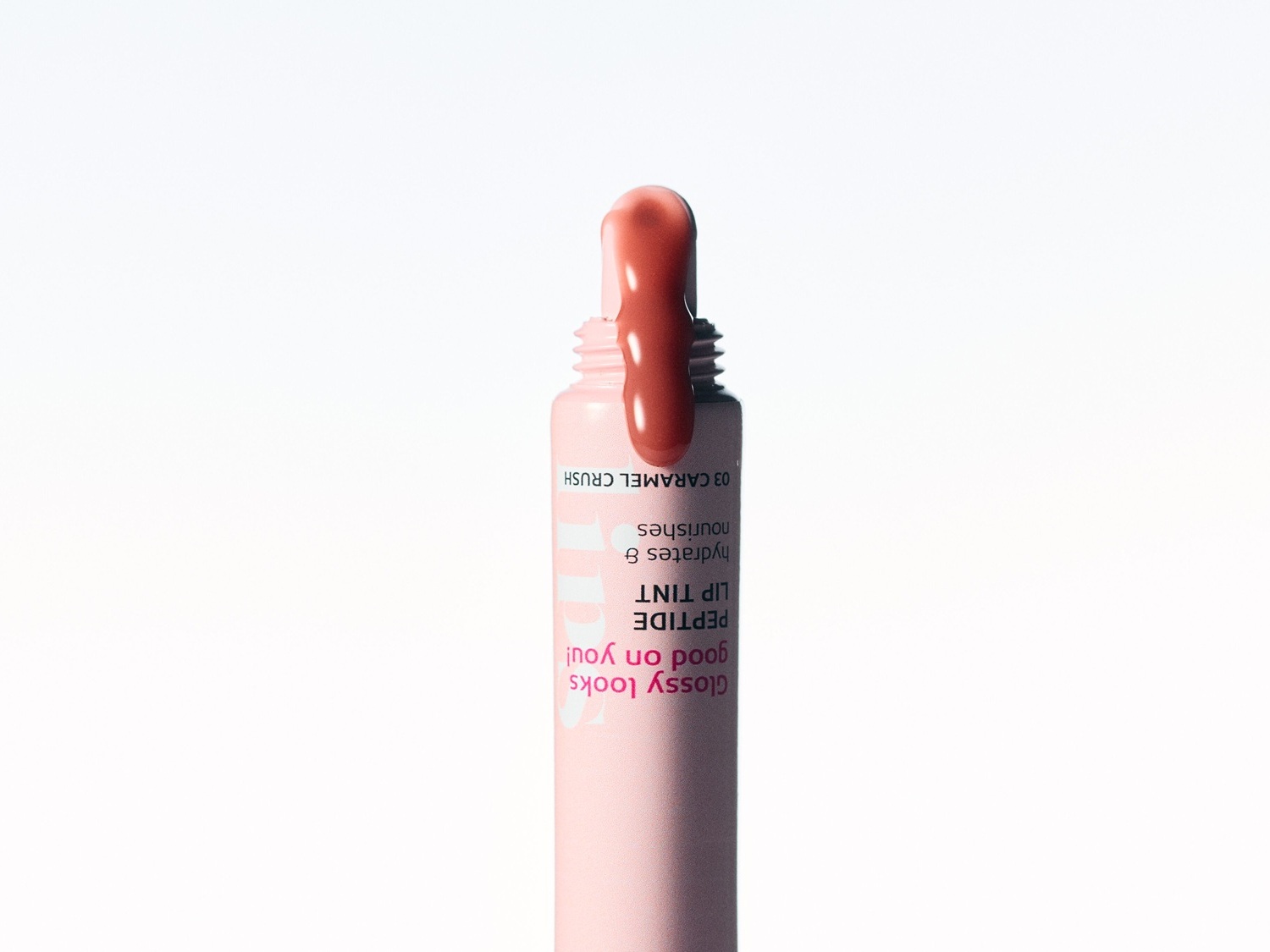 Das Bild zeigt einen geöffneten Lipgloss in der Farbe 03 Caramel Crush, der als Peptid-Lippentönung beschrieben wird, die Feuchtigkeit spendet und pflegt, mit dem Slogan "Glossy looks good on you!" auf der rosa Verpackung.