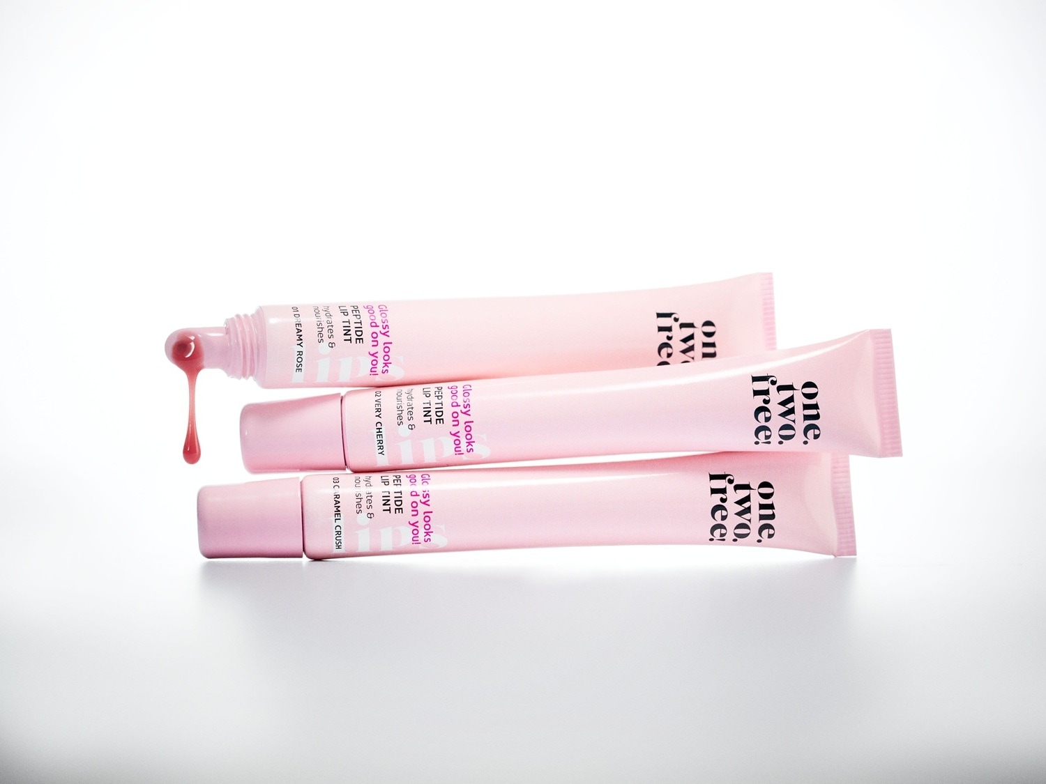 Drei Tuben des One.Two.Free! Peptide Lip Tint in den Farben Dreamy Rose, Very Cherry und Caramel Crush, wobei aus der oberen Tube ein Tropfen des rosafarbenen Lip Tints herausquillt, der Feuchtigkeit spendet und pflegt.