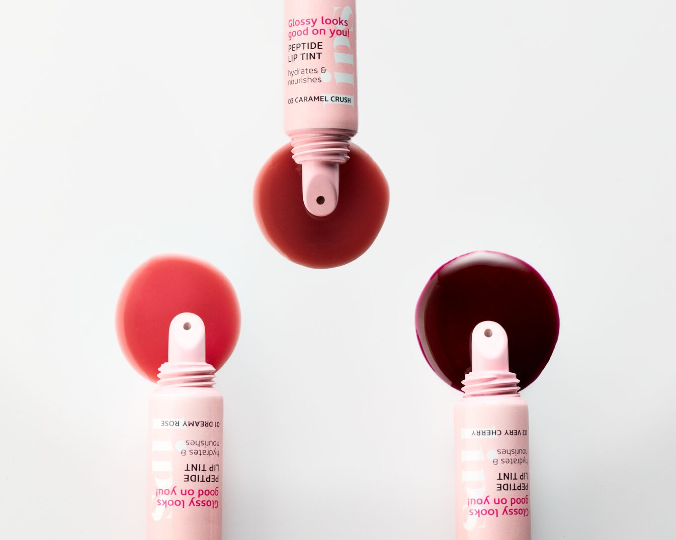 Drei offene Lip-Tint-Tuben in Rosatönen, die jeweils eine glänzende, flüssige Lippenfarbe in den Nuancen Karamell-Crush, Dreamy Rose und Very Cherry zeigen, die Feuchtigkeit spendet und pflegt.