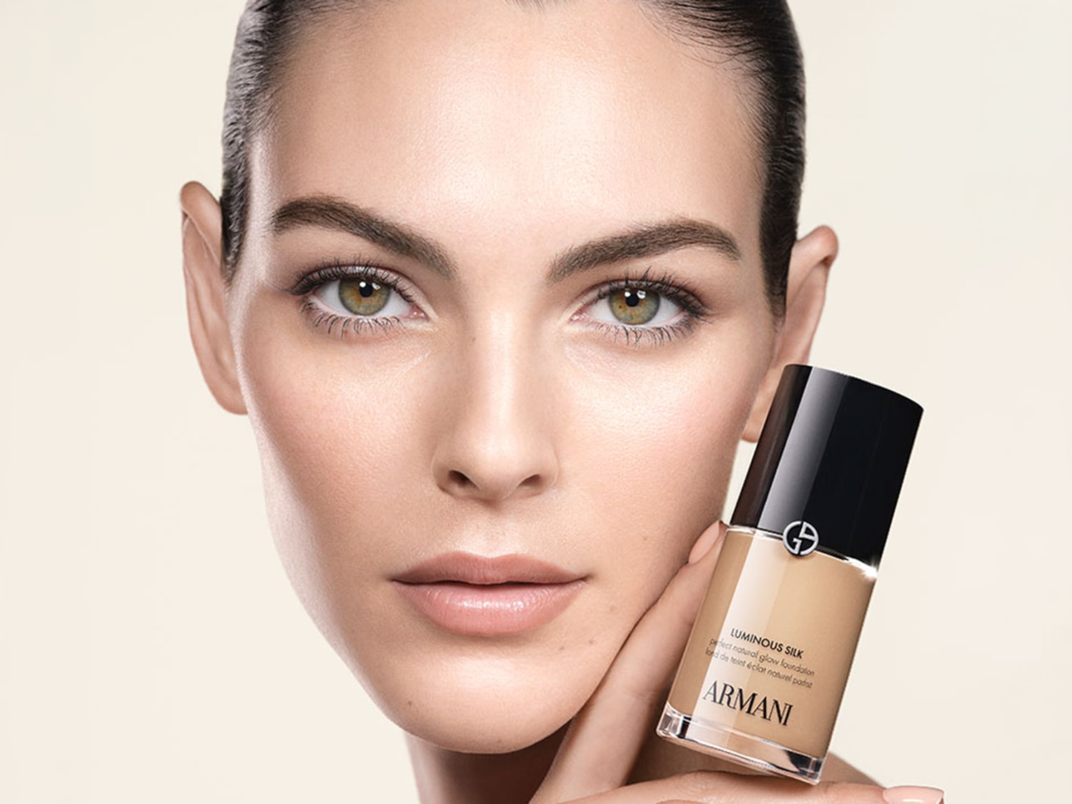 Prim-plan al unei femei cu ten luminos și machiaj natural, ținând în mână un flacon de fond de ten Armani, pe fundal deschis