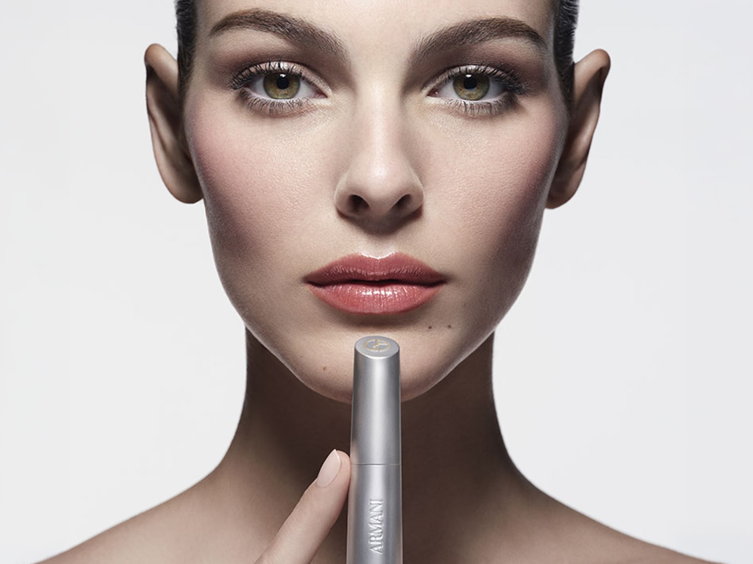 Prim-plan al unei femei cu machiaj elegant în nuanțe neutre, ținând în fața bărbiei produsul de buze Armani beauty Prisma Flash pe fundal gri deschis.