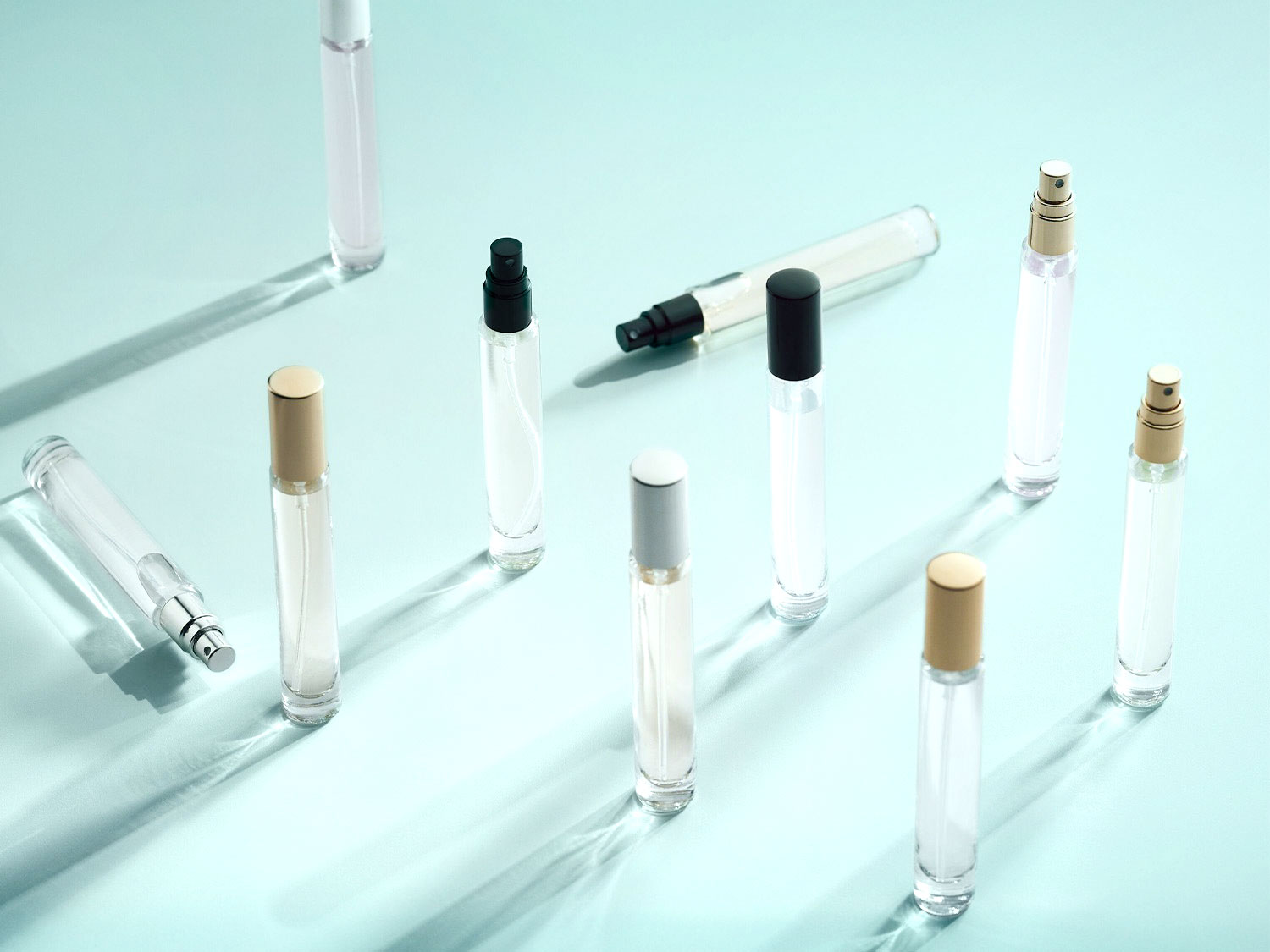 Plusieurs flacons de parfum transparents avec des bouchons dorés, argentés et noirs sont disposés sur une surface bleu clair, créant un motif élégant et moderne.