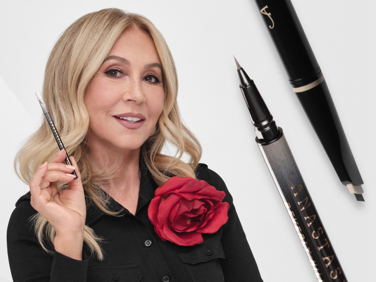 Anastasia Soare, fondatrice d'Anastasia Beverly Hills, tient un crayon à sourcils noir et blanc, vêtue d'une chemise noire ornée d'une rose rouge, avec deux autres produits pour les sourcils en arrière-plan.