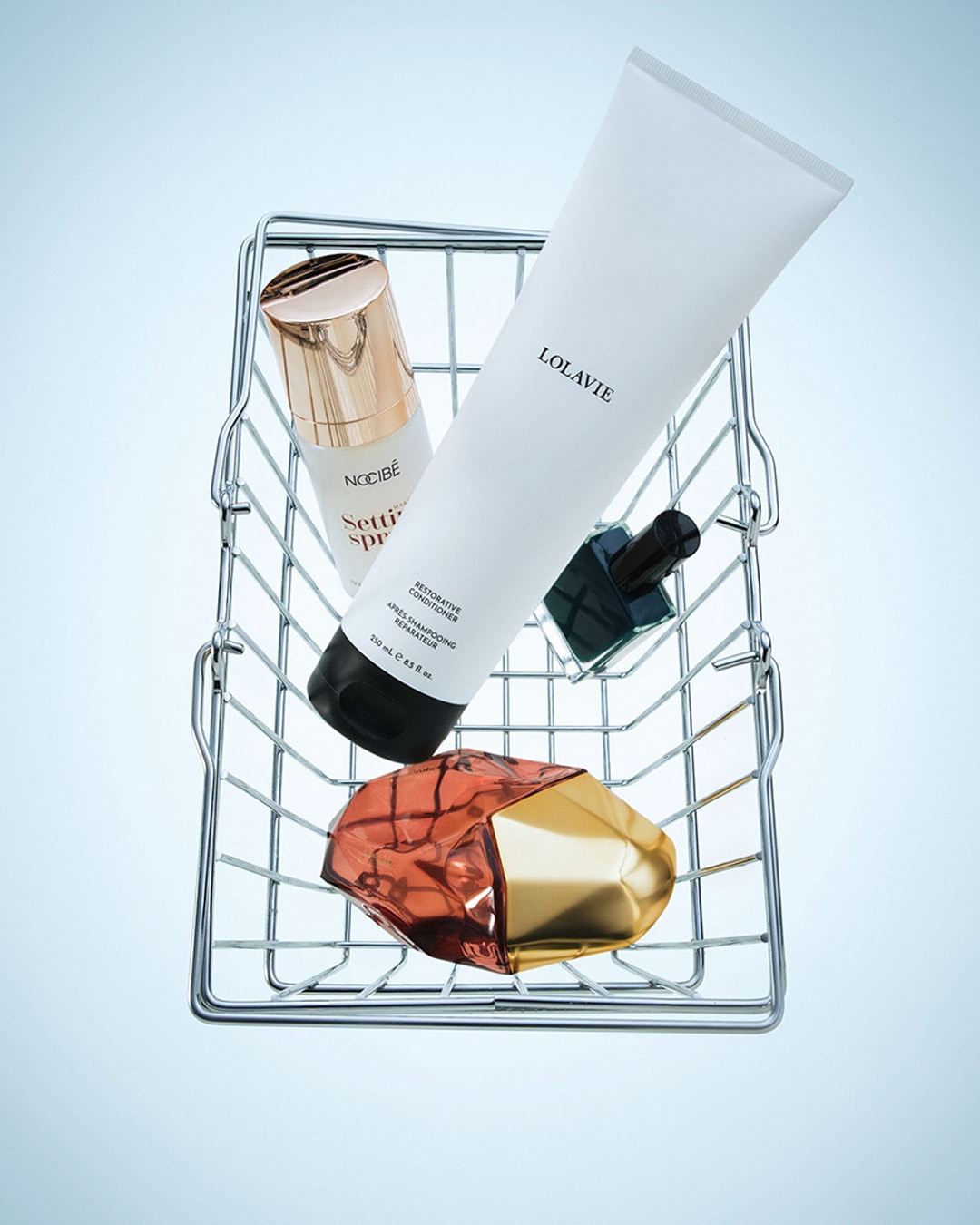 Un panier en fil de fer contient un vaporisateur fixant blanc et or de Nocibé, un après-shampooing réparateur blanc de Lolavie, un petit flacon de vernis à ongles noir et un flacon de parfum doré et cuivré.