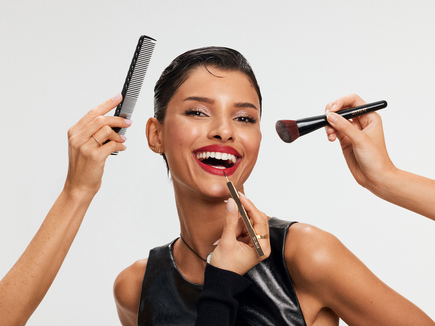 Une femme souriante aux cheveux foncés et à la peau bronzée se fait maquiller les lèvres en rouge vif, tandis que des mains tiennent un peigne noir et un pinceau de maquillage autour d'elle.