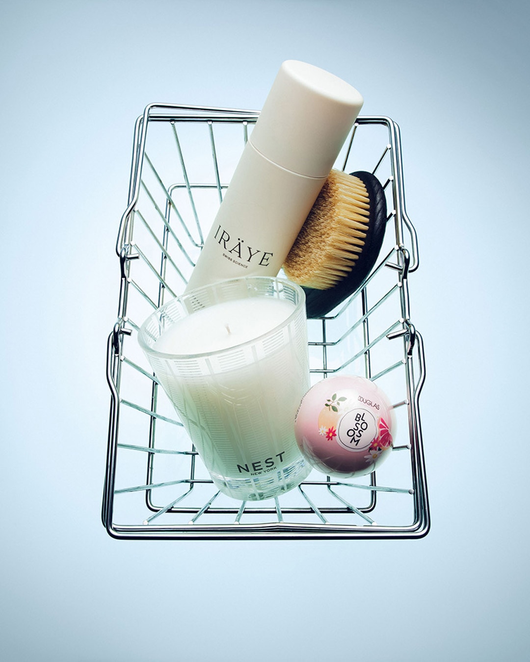 Un panier en fil de fer contient une bouteille blanche d'IRÄYE Swiss Science, une brosse de bain en bois, une bougie parfumée Nest New York et une bombe de bain rose Douglas Blossom.