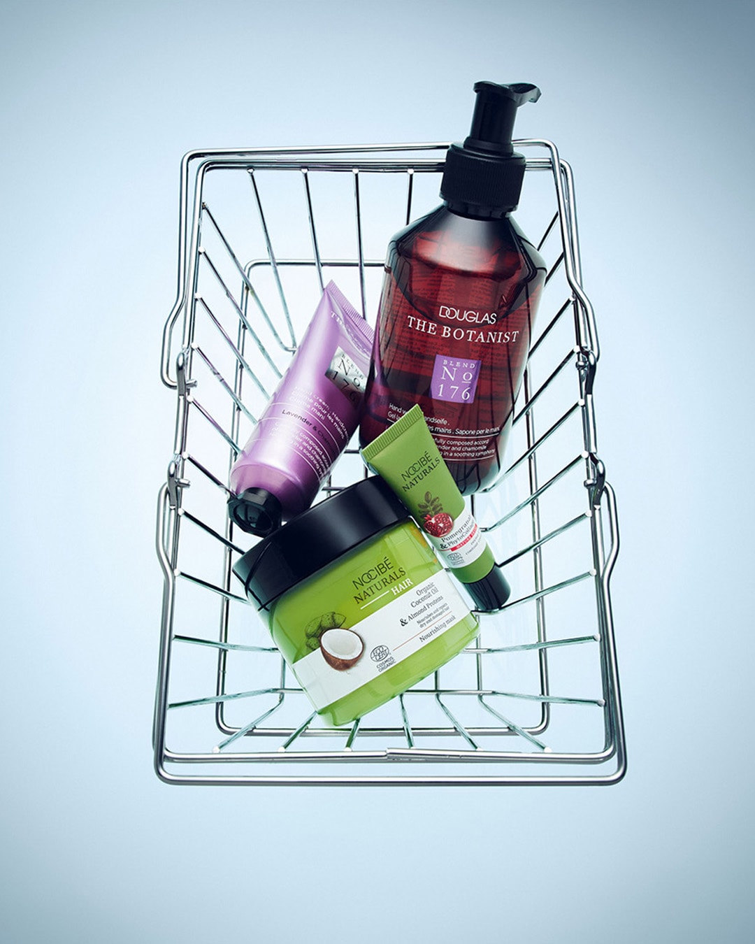 Un panier en fil de fer contient quatre produits de beauté : un flacon de savon liquide Douglas The Botanist Blend No 176, un tube de crème pour les mains à la lavande, un tube de crème à la grenade et au thym, et un pot de masque capillaire Nocibé Naturals à l'huile de coco et aux protéines d'amande.