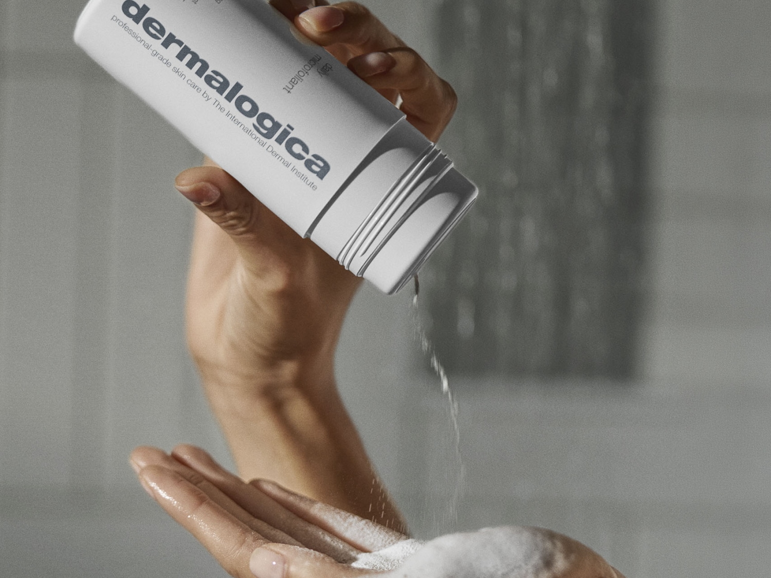 Une main tient une bouteille blanche de Dermalogica Daily Microfoliant, versant la poudre exfoliante dans la paume de l'autre main, où elle se transforme en une mousse blanche et légère.