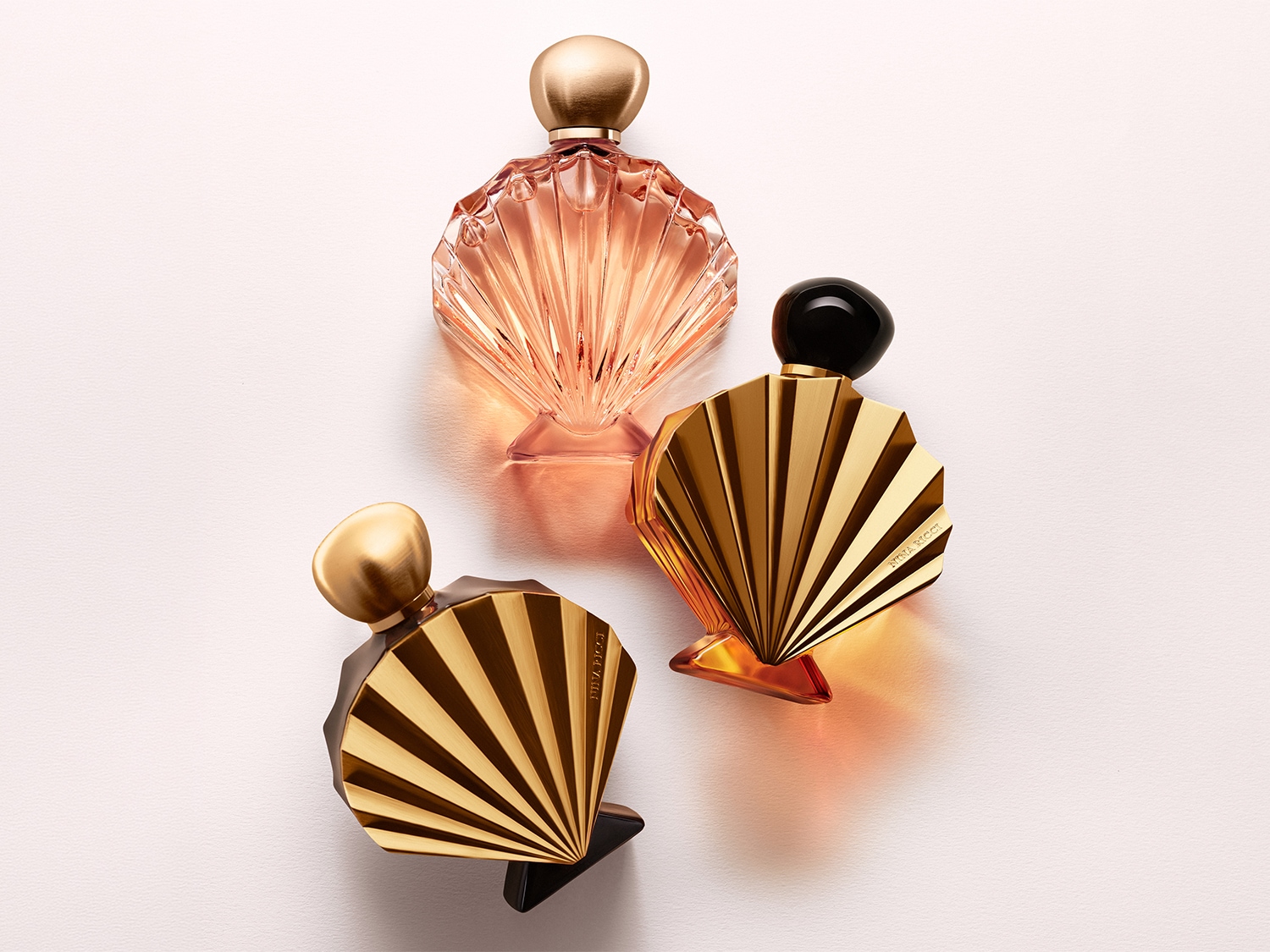 Trois flacons de parfum en forme d'éventail, un rose et deux dorés, sont disposés sur une surface blanche, capturant l'élégance et le luxe.