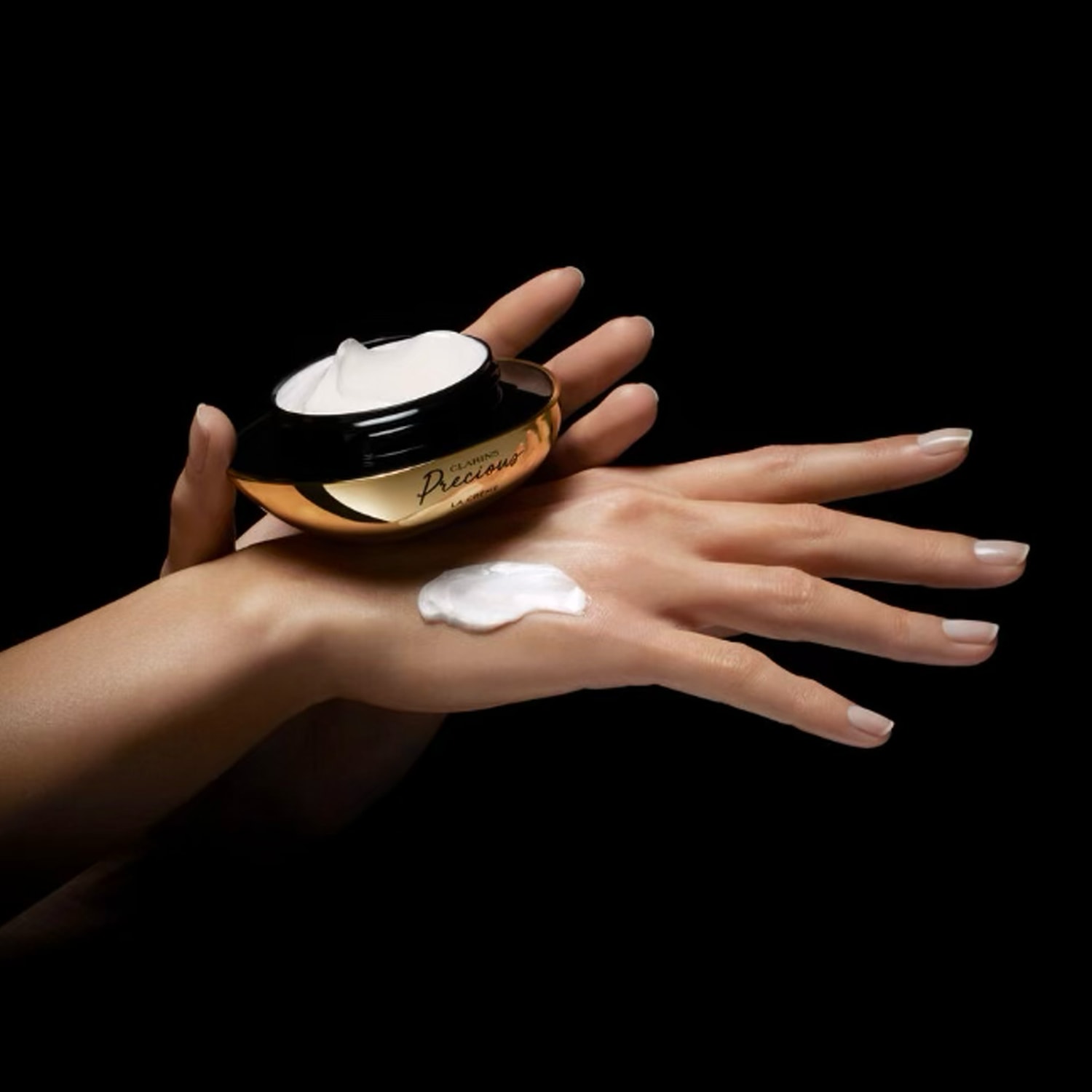 Une main tient un pot de crème Clarins Precious La Crème noir et or, tandis qu'une autre main présente une noisette de crème blanche, illustrant la texture riche du produit.