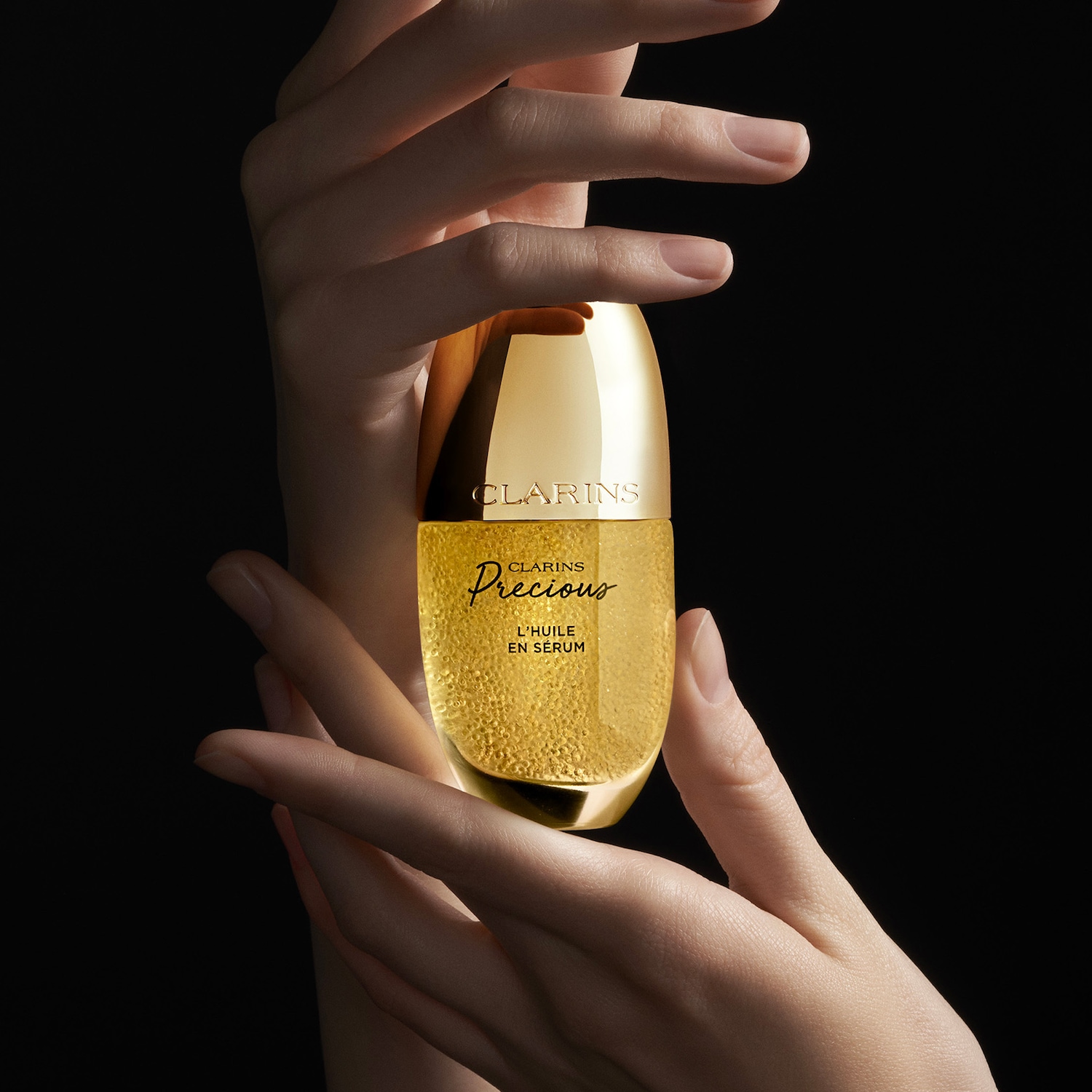 Deux mains tiennent délicatement le sérum Clarins Precious L'Huile en Sérum, un flacon doré élégant rempli d'un liquide doré scintillant, sur un fond noir.