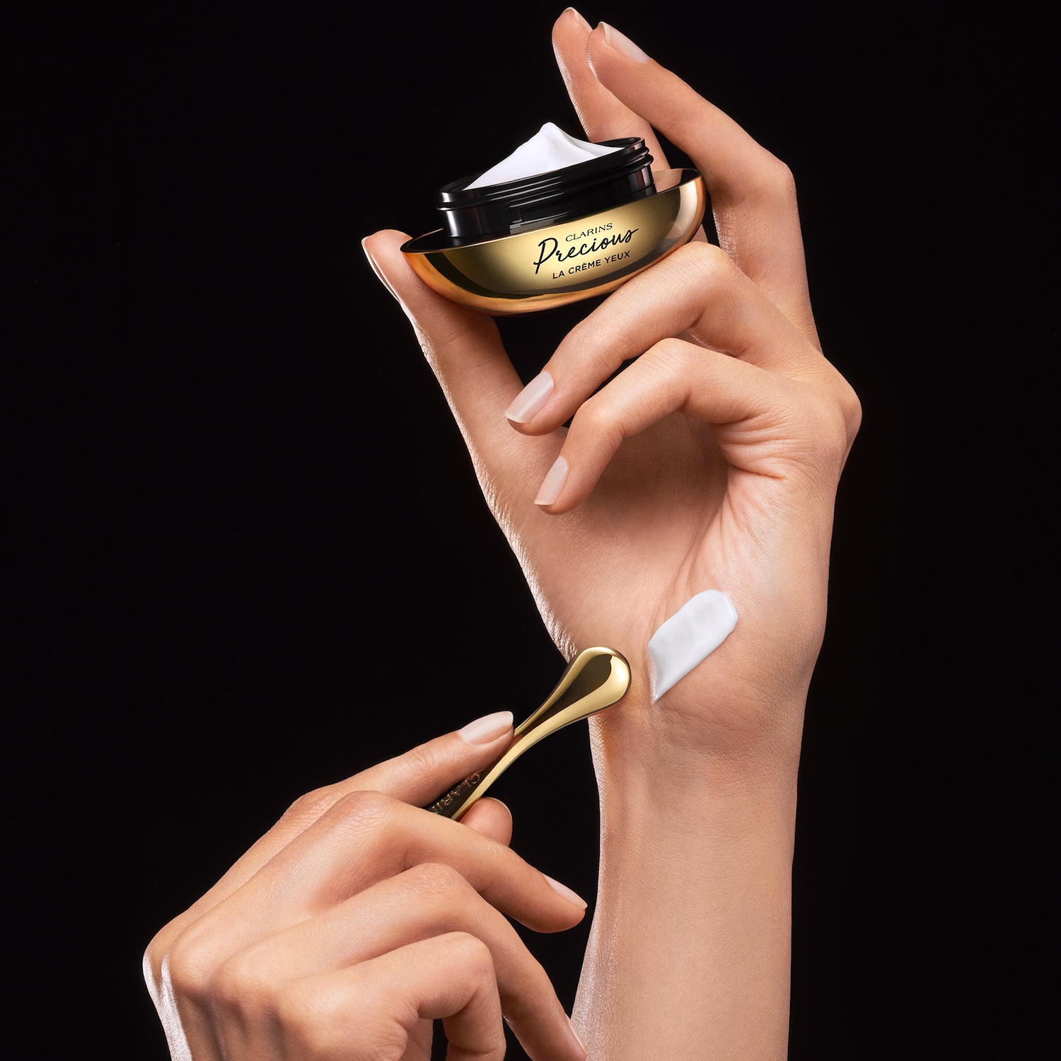 Une main tient un pot de crème pour les yeux Clarins Precious La Crème Yeux doré et noir, tandis qu'une autre main applique une petite quantité de crème blanche sur le dos de la main à l'aide d'une spatule dorée.