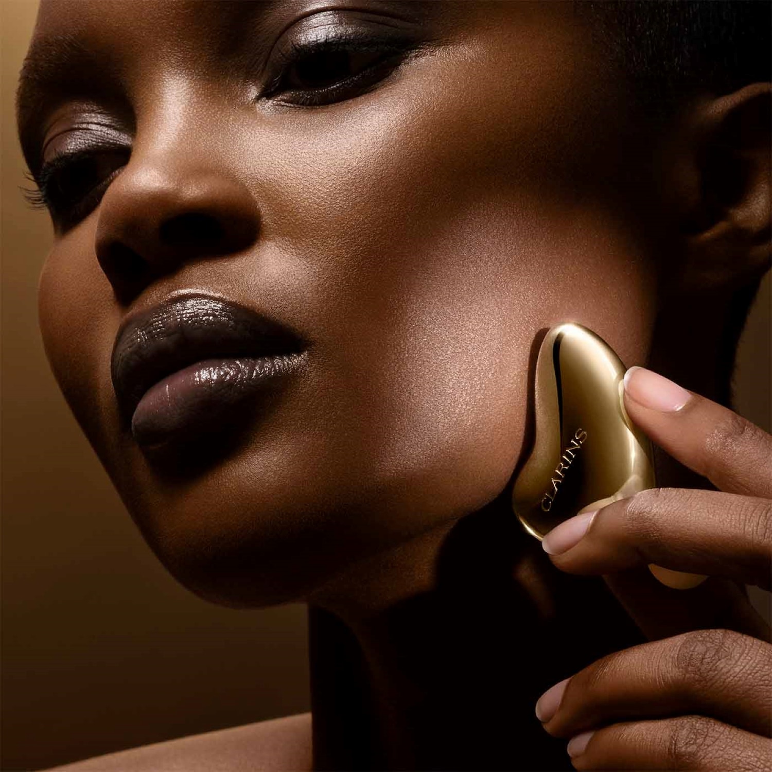 Une femme à la peau foncée utilise un outil de massage facial doré de Clarins sur sa joue, avec un maquillage des yeux charbonneux et des lèvres brillantes.