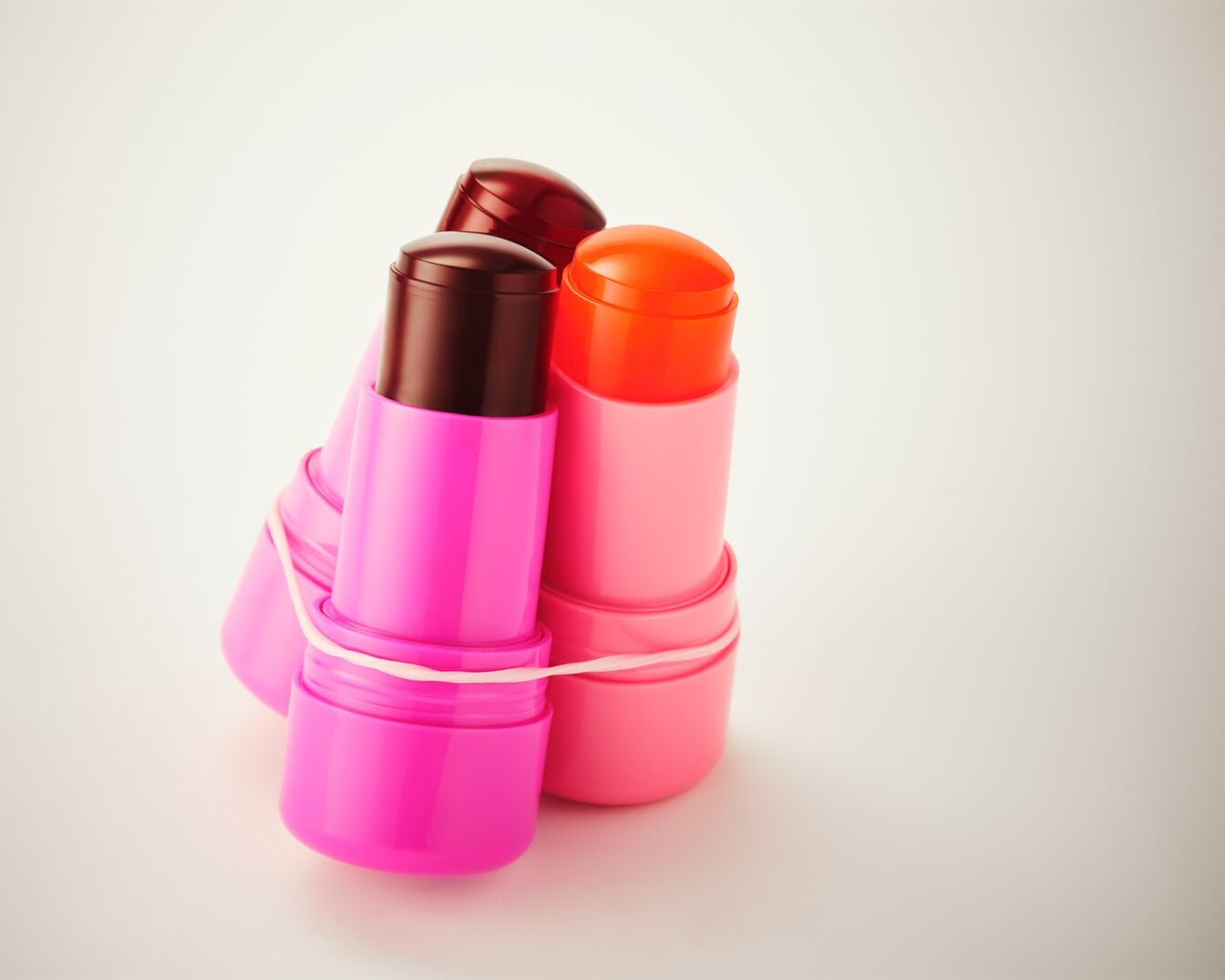 Drei Lippenstifte in leuchtendem Pink, Hellrosa und Dunkelrot, zusammengehalten von einem weißen Gummiband, stehen aufrecht vor einem hellen Hintergrund.