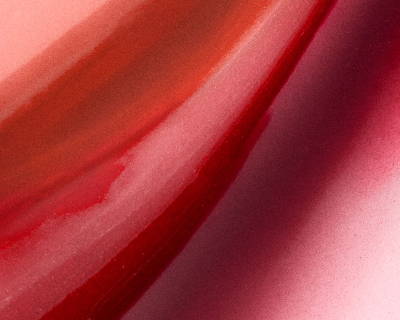 Ein Nahaufnahme-Bild zeigt drei glänzende, flüssige Lippenstift-Swatches in verschiedenen Rottönen, von hellem Korallenrot über tiefes Burgunderrot bis hin zu einem satten Himbeerrot, die diagonal über das Bild verlaufen.