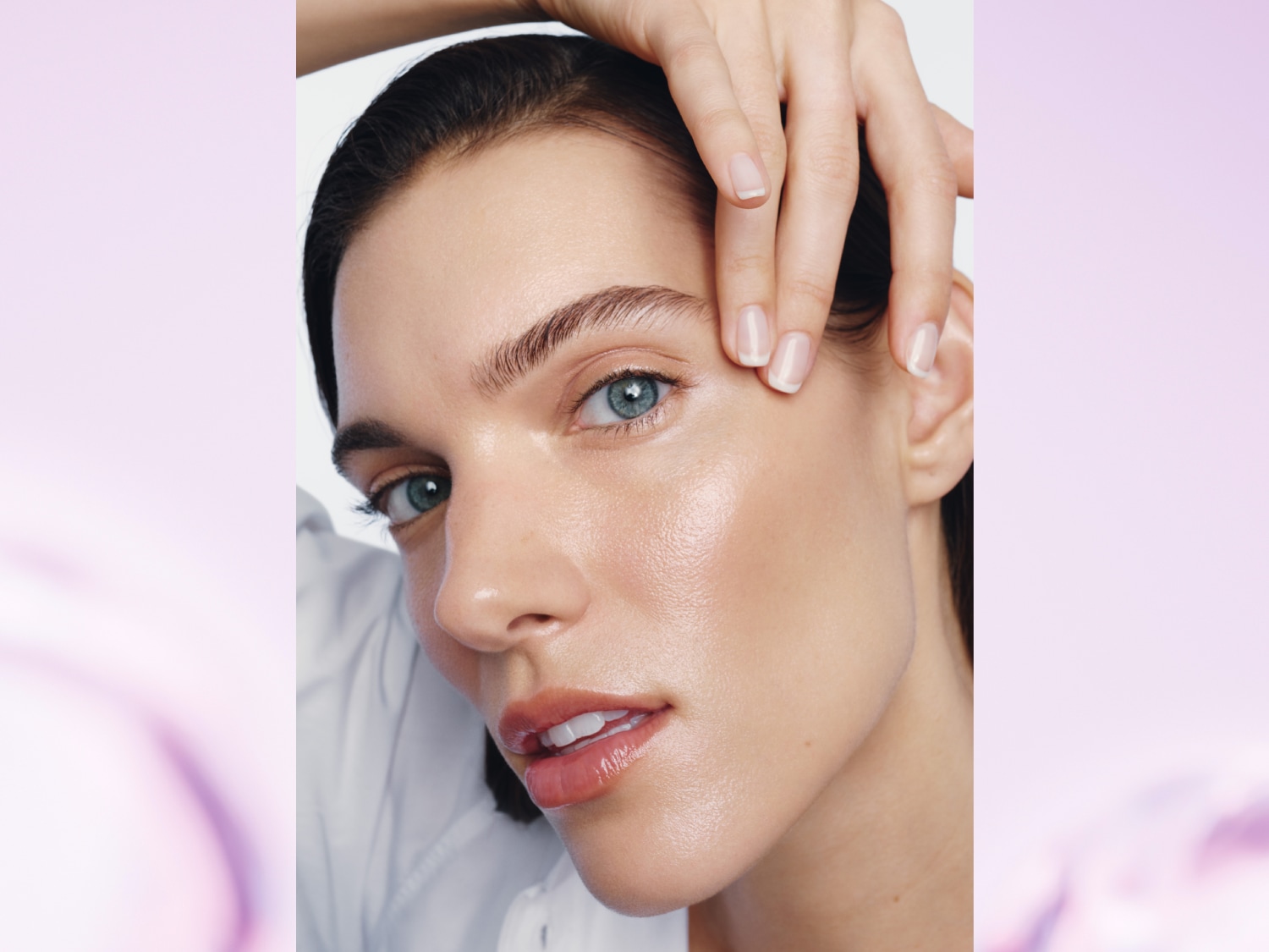 Une femme aux cheveux foncés et aux yeux bleus, vêtue d'une chemise blanche, touche son visage d'une main aux ongles soignés, mettant en valeur sa peau éclatante et ses lèvres brillantes.