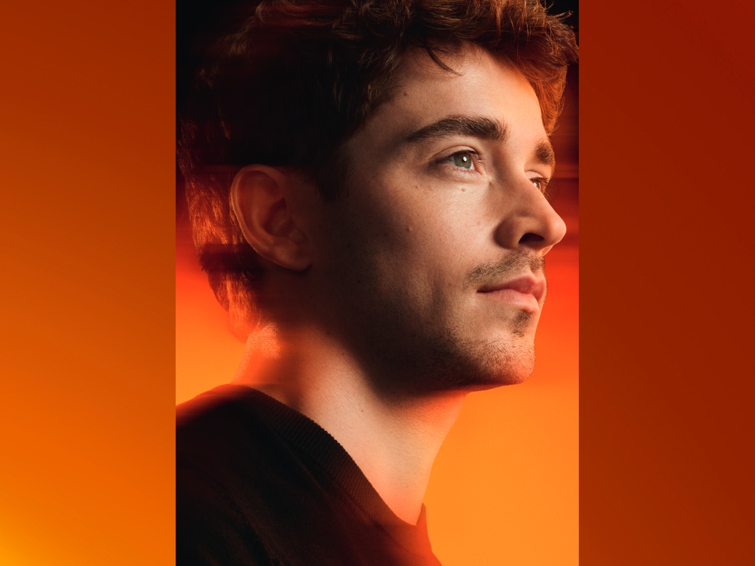 Un homme aux cheveux bruns et aux yeux verts, vêtu d'un t-shirt noir, regarde vers la droite sur un fond orange vif.