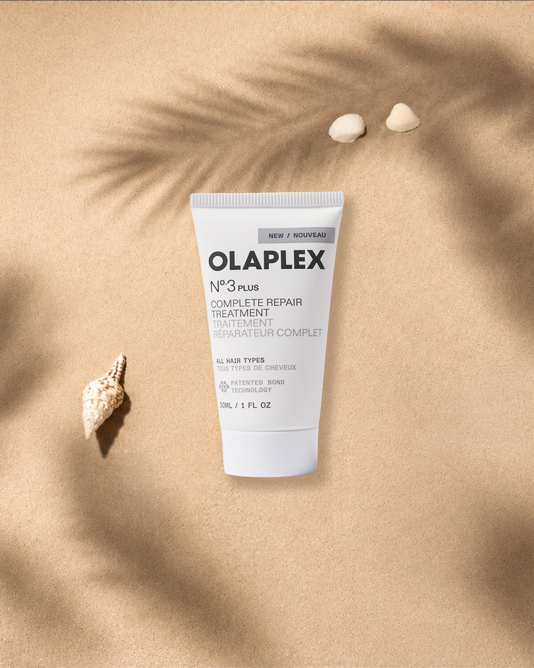 Die Olaplex N°3 Plus Complete Repair Treatment, eine weiße Tube mit schwarzer Schrift, liegt auf sandfarbenem Untergrund, umgeben von Muscheln und Palmenblattschatten, und ist für alle Haartypen geeignet.
