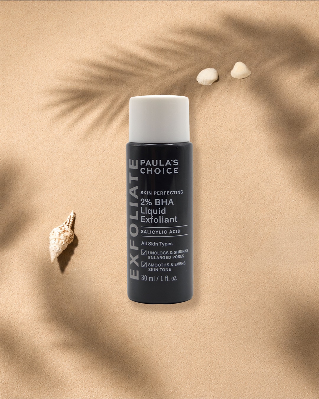 Die Paula's Choice 2% BHA Liquid Exfoliant Flasche in Schwarz mit weißem Deckel steht auf sandigem Untergrund, umgeben von Muscheln und Palmenblattschatten, und verspricht, Poren zu reinigen und das Hautbild zu glätten.