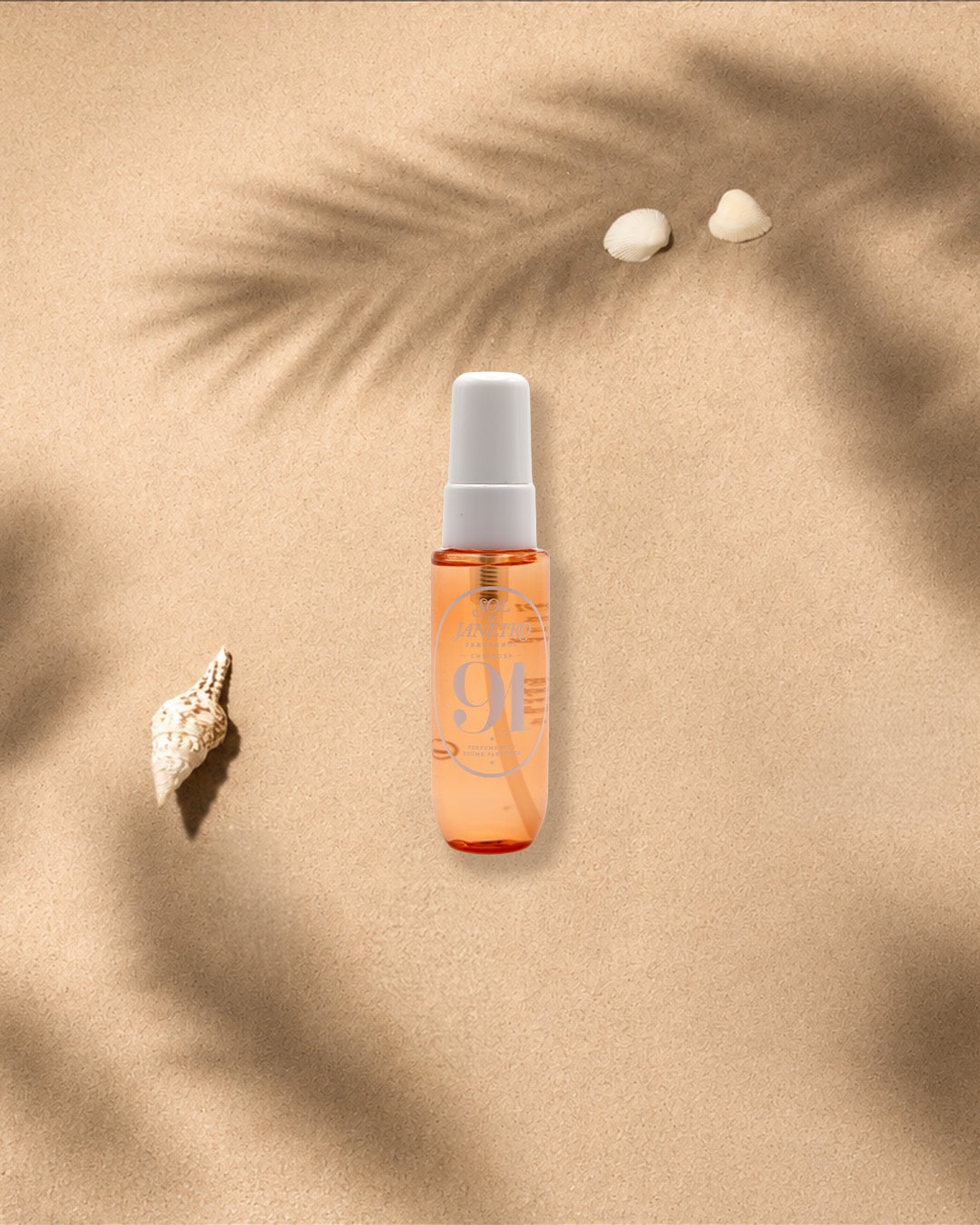 Ein orangefarbenes Parfümspray mit weißem Deckel steht auf beigem Sand, umgeben von Muscheln und Palmenblattschatten, die eine sommerliche Atmosphäre schaffen.