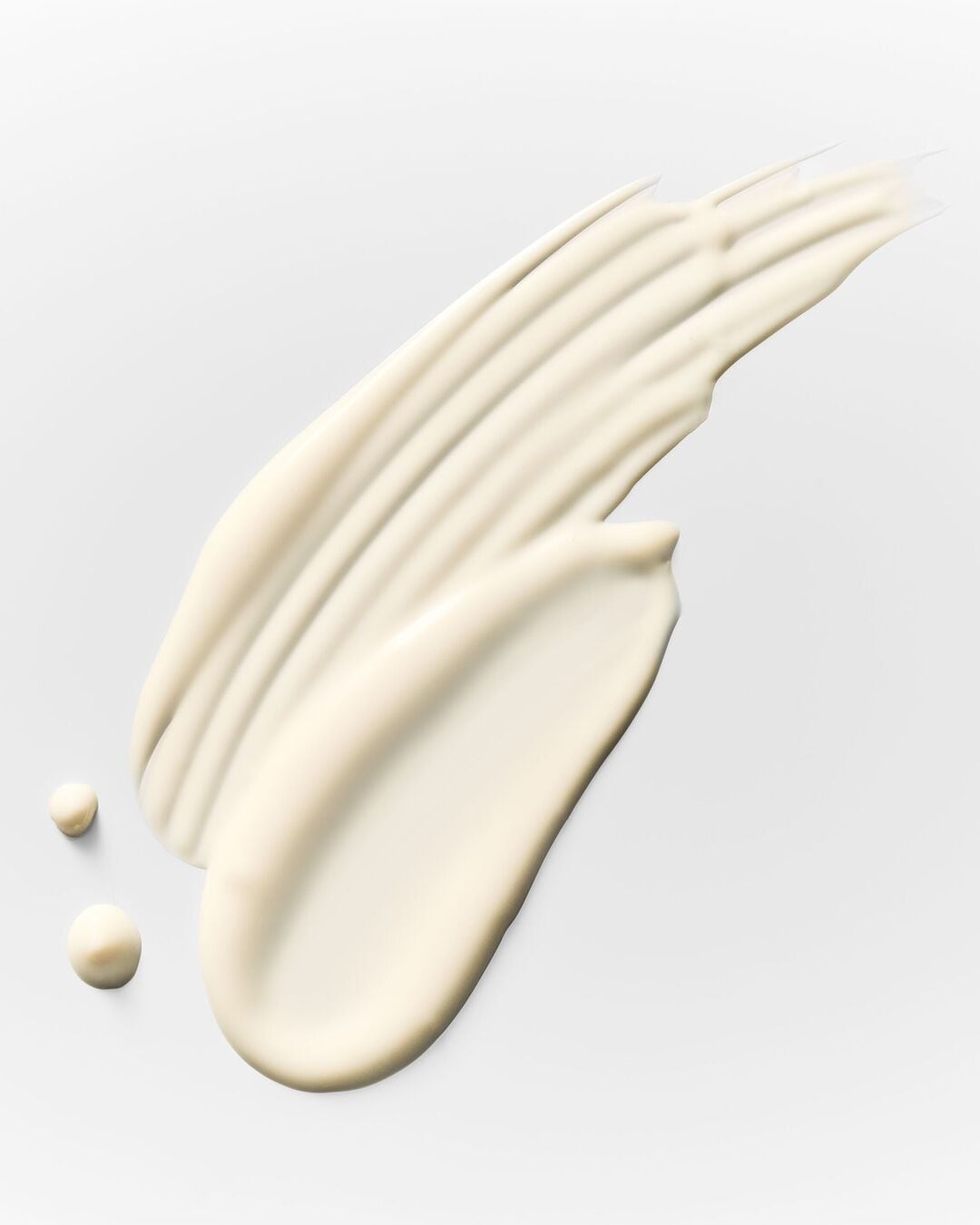 Une texture crémeuse et riche, de couleur beige clair, est étalée en un mouvement fluide, avec deux petites gouttes de crème à côté, sur un fond blanc uni.