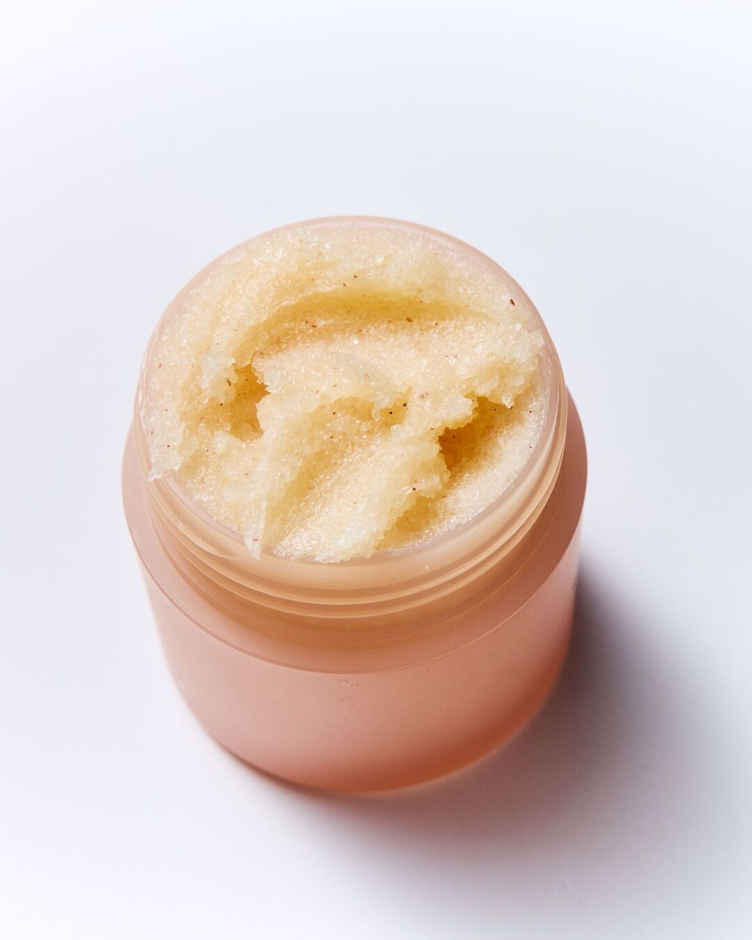 Un pot ouvert de gommage pour les lèvres, de couleur pêche, révèle une texture granuleuse et hydratante, parfaite pour exfolier et adoucir les lèvres.