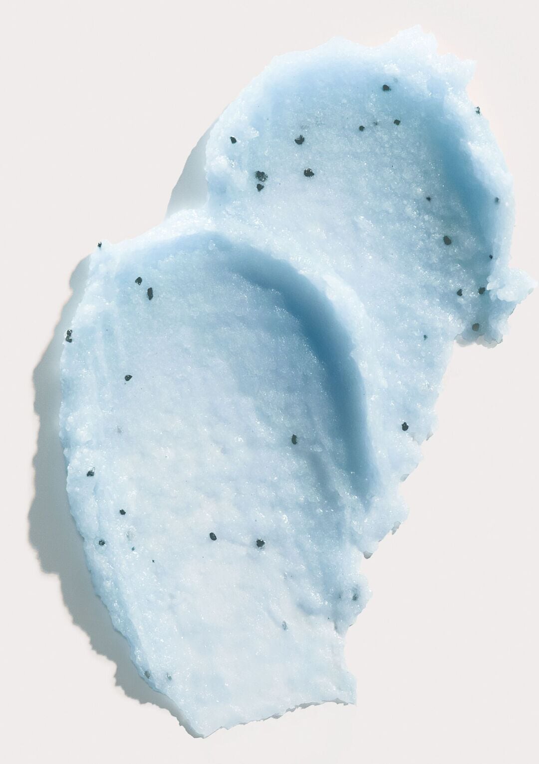 Un gros plan sur un gommage corporel bleu clair avec de petits grains exfoliants noirs, étalé sur une surface blanche, créant une texture riche et invitante.