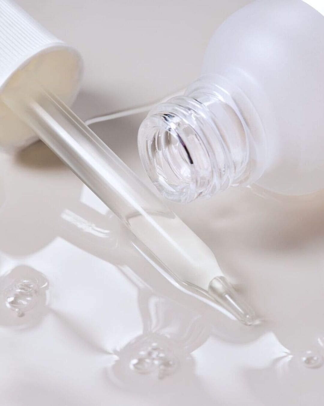 Un flacon de sérum blanc mat est posé sur le côté, avec son compte-gouttes en verre transparent à côté, tous deux entourés de gouttes de sérum blanc laiteux.