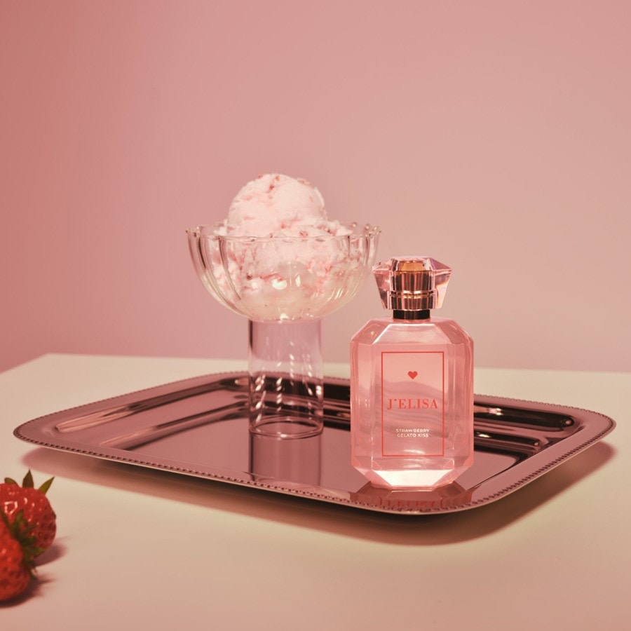 Ein rosafarbener Parfümflakon von J'ELISA mit der Aufschrift "STRAWBERRY GELATO KISS" steht auf einem silbernen Tablett neben einer Glasschale mit Erdbeereis und frischen Erdbeeren, alles vor einem rosafarbenen Hintergrund.