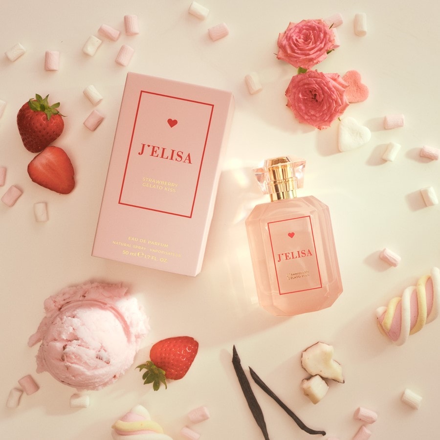 Das J'ELISA Strawberry Gelato Kiss Eau de Parfum ist in einer rosafarbenen Schachtel verpackt, die mit einem roten Herz und dem Namen des Parfums verziert ist, umgeben von frischen Erdbeeren und kleinen Marshmallows.