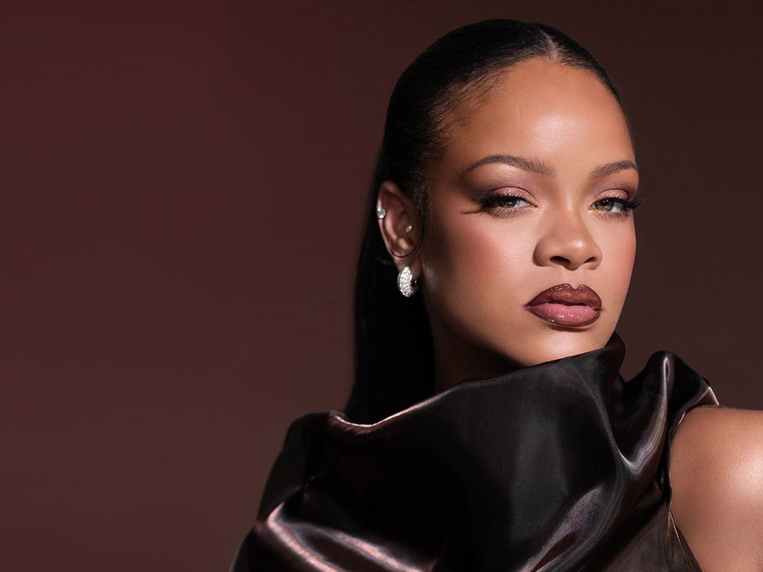Rihanna trägt ein glänzendes, dunkles Oberteil und hat ihr Haar zu einem eleganten Pferdeschwanz zurückgebunden, während sie mit einem intensiven Blick in die Kamera schaut.