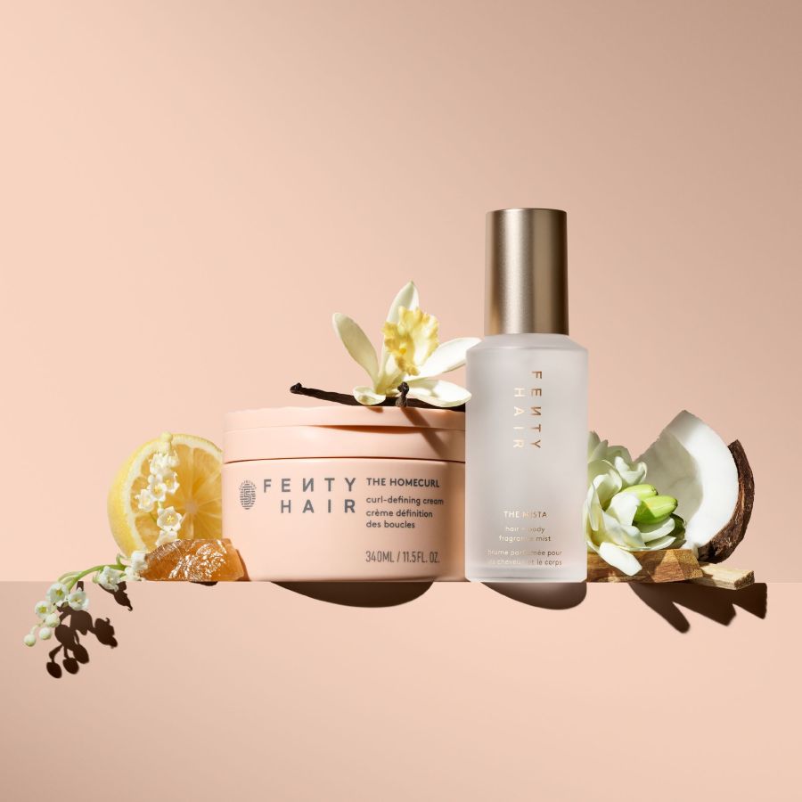 Fenty Hair The Homecurl lockendefinierende Creme in einem pfirsichfarbenen Tiegel und The Mista Haar- und Körperduftnebel in einer milchig-weißen Flasche, umgeben von Vanilleblüten, Zitronen- und Kokosnussstücken.