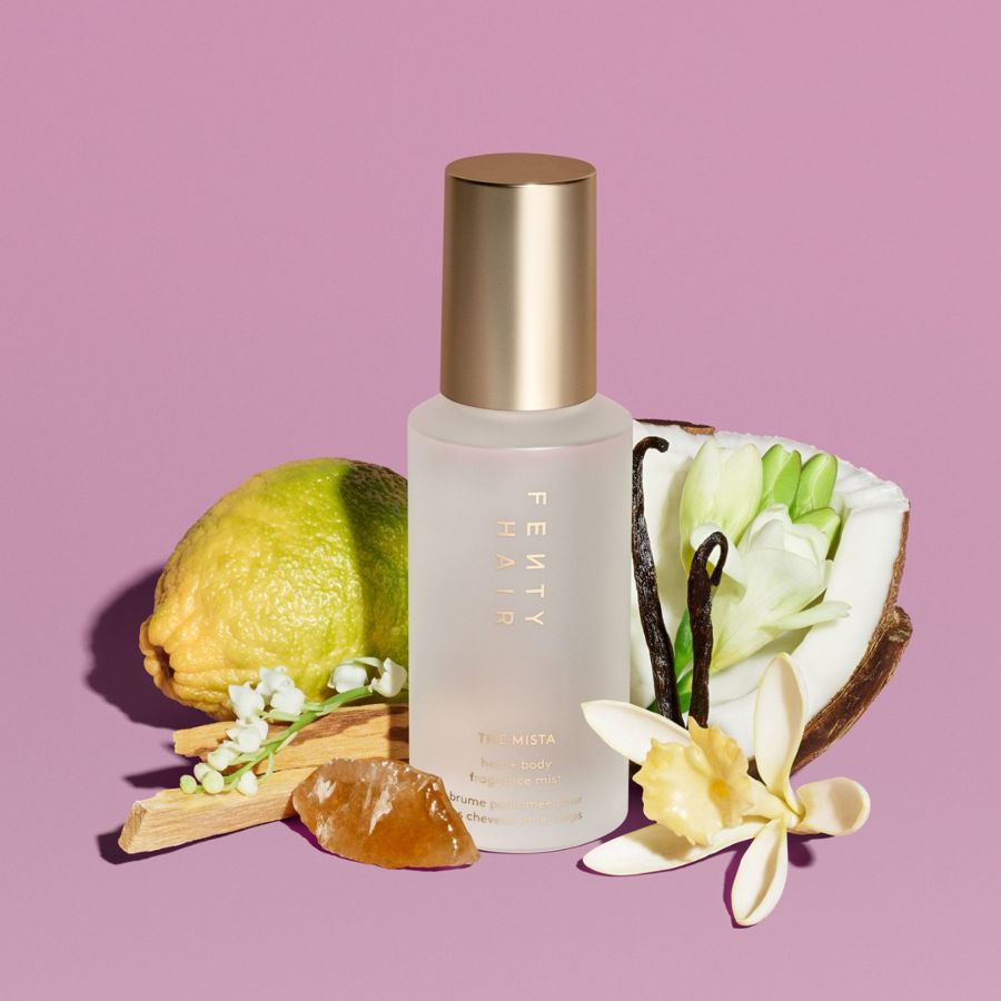 Fenty Hair The Mista Hair & Body Fragrance Mist in einer durchsichtigen Flasche mit goldener Kappe, umgeben von Limette, Kokosnuss, Vanilleschoten und Blumen auf lila Hintergrund.