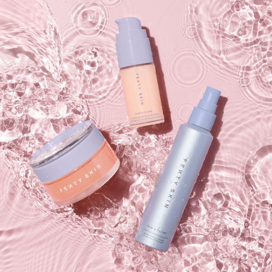 Trois produits Fenty Skin, dont une crème hydratante, un sérum et une brume pour le visage, sont présentés sur un fond rose clair avec des ondulations d'eau.