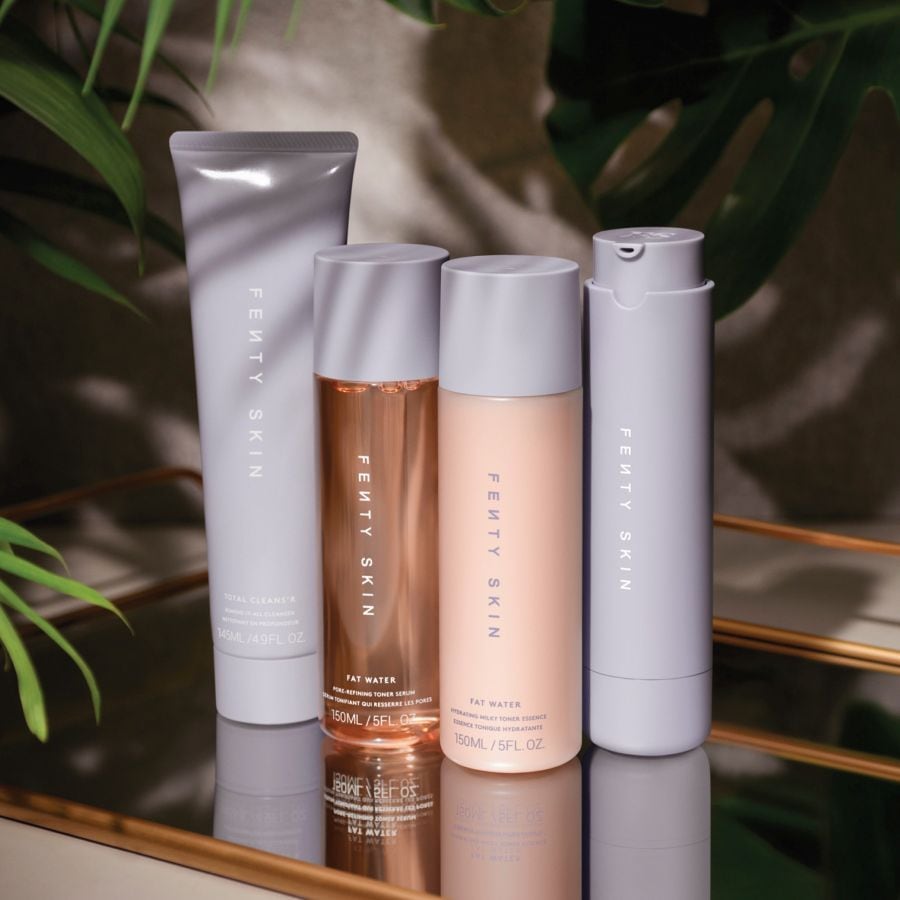 Découvrez la gamme Fenty Skin, comprenant un nettoyant, un sérum tonique affinant les pores et une essence tonique hydratante, présentés sur un plateau doré avec des feuilles vertes en arrière-plan.