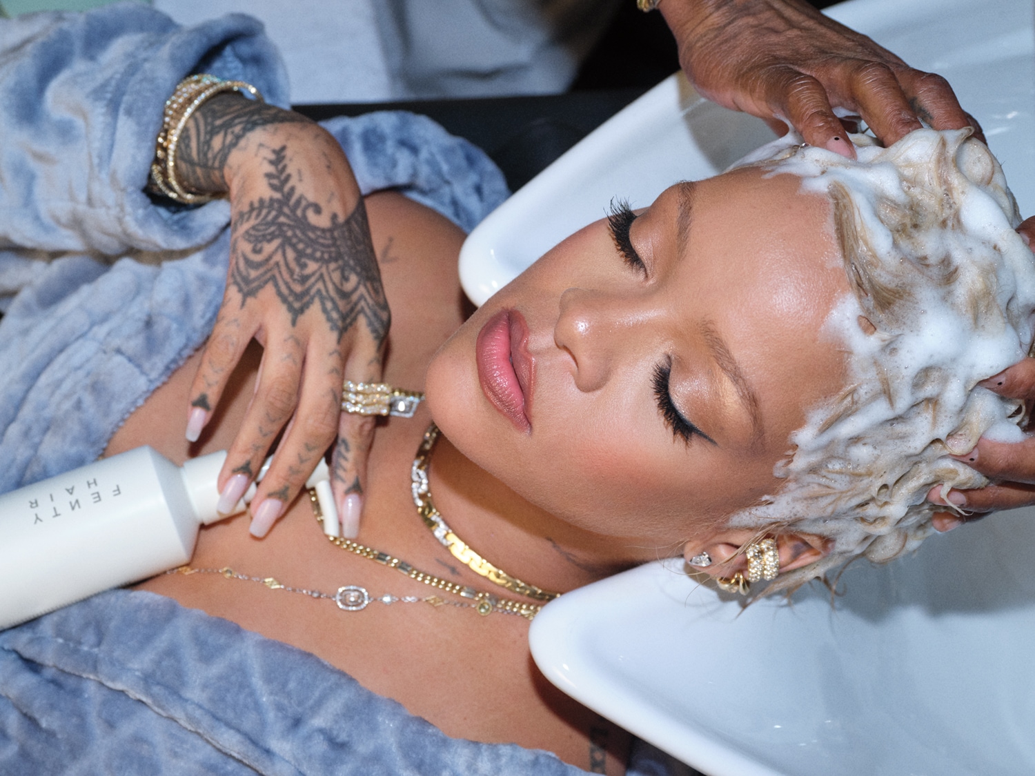 Une femme aux cheveux blonds se fait laver les cheveux avec un shampoing moussant, tandis qu'elle tient une bouteille blanche de Fenty Hair, vêtue d'une robe de chambre bleue et de bijoux en or.