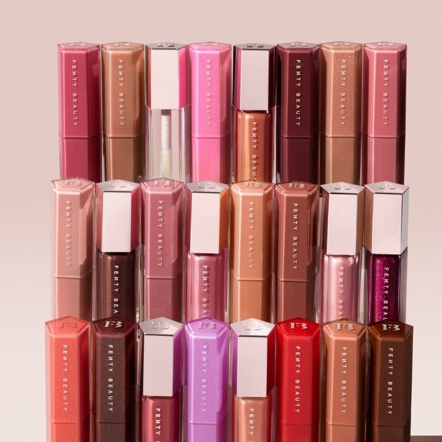 Une collection de brillants à lèvres Fenty Beauty est présentée, avec des tubes hexagonaux de différentes nuances de rose, de nude et de marron, certains avec des finitions scintillantes, disposés en rangées ordonnées.