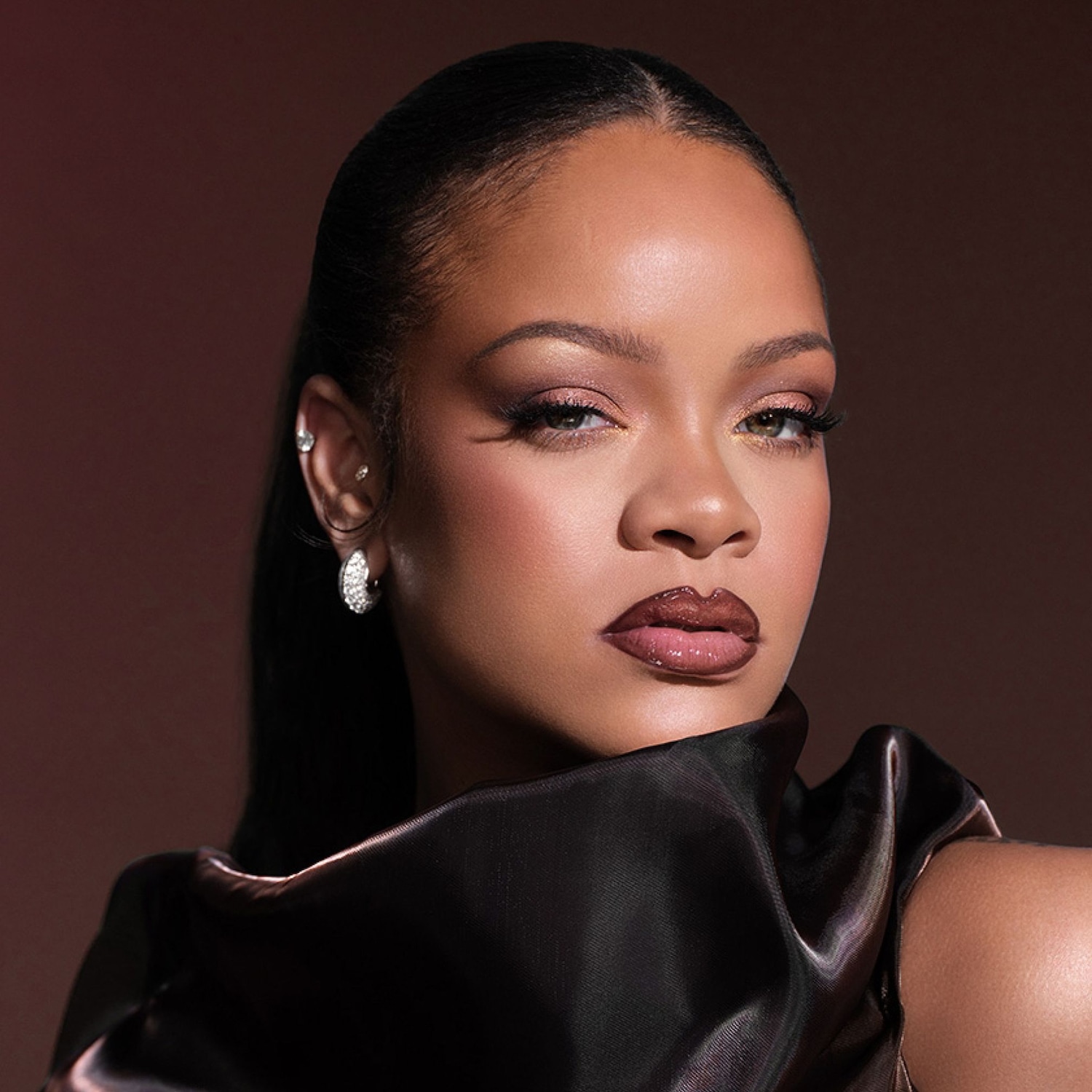 Rihanna porte un maquillage glamour avec des yeux charbonneux, des lèvres brillantes et un teint éclatant, mettant en valeur des boucles d'oreilles scintillantes et un haut en satin noir.
