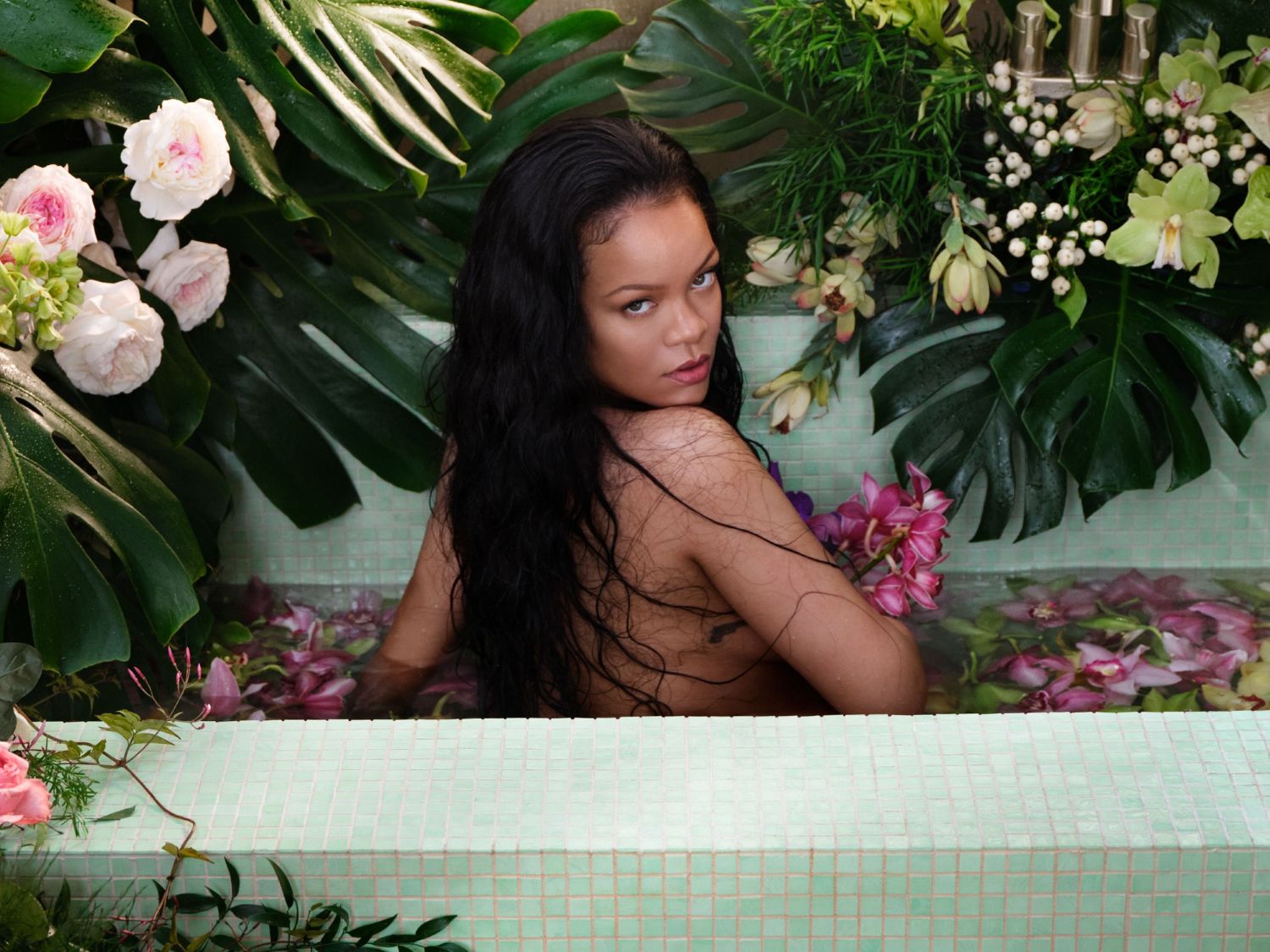 Rihanna est allongée dans une baignoire carrelée de couleur menthe, remplie d'eau et de pétales de fleurs roses et violettes, entourée de grandes feuilles vertes et de fleurs blanches et roses.